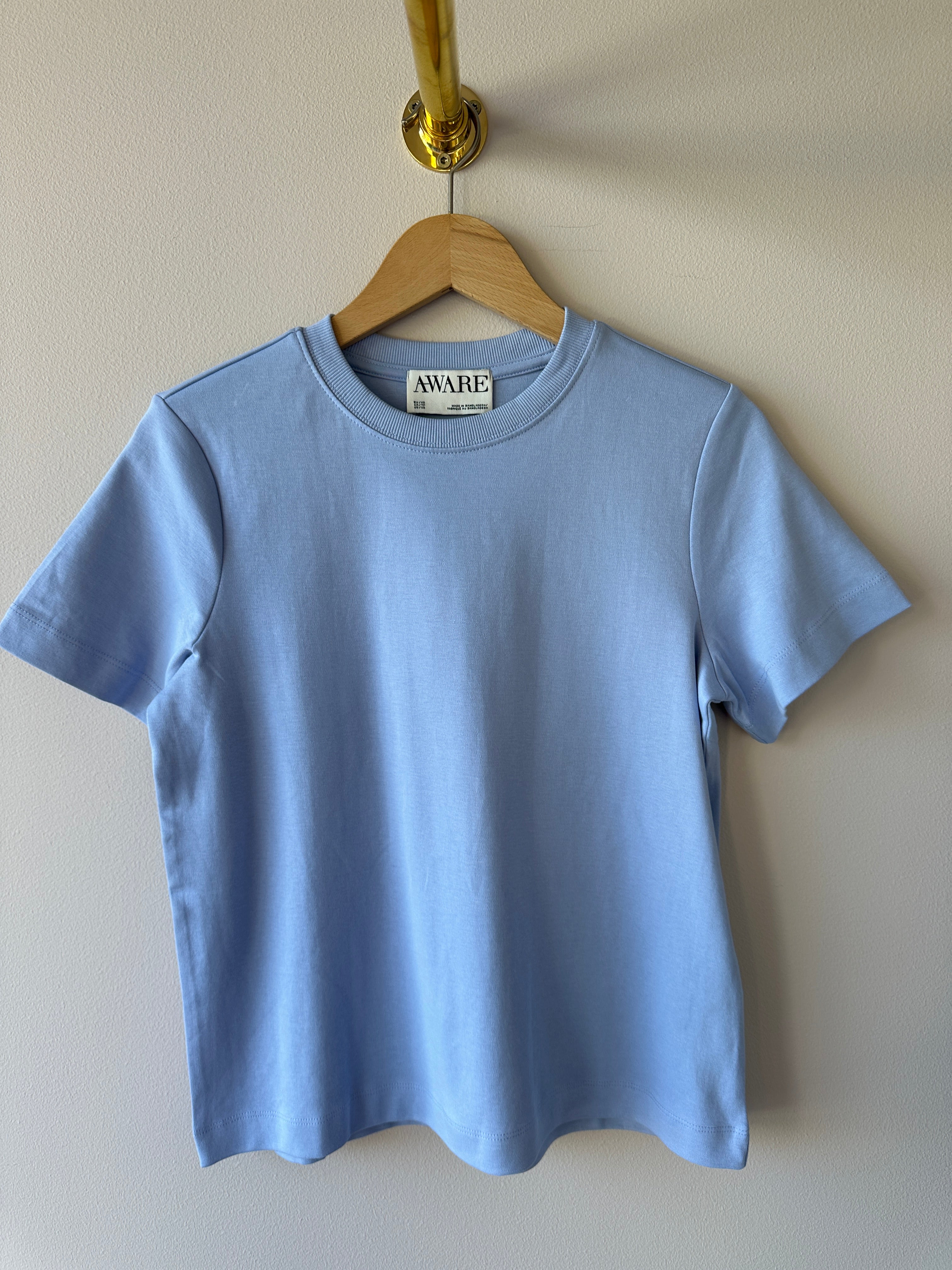 Ljusblå t-shirt med rund ribbad hals och korta ärmar hängande på galge mot vit vägg, etikett med texten AWARE synlig i halsen.