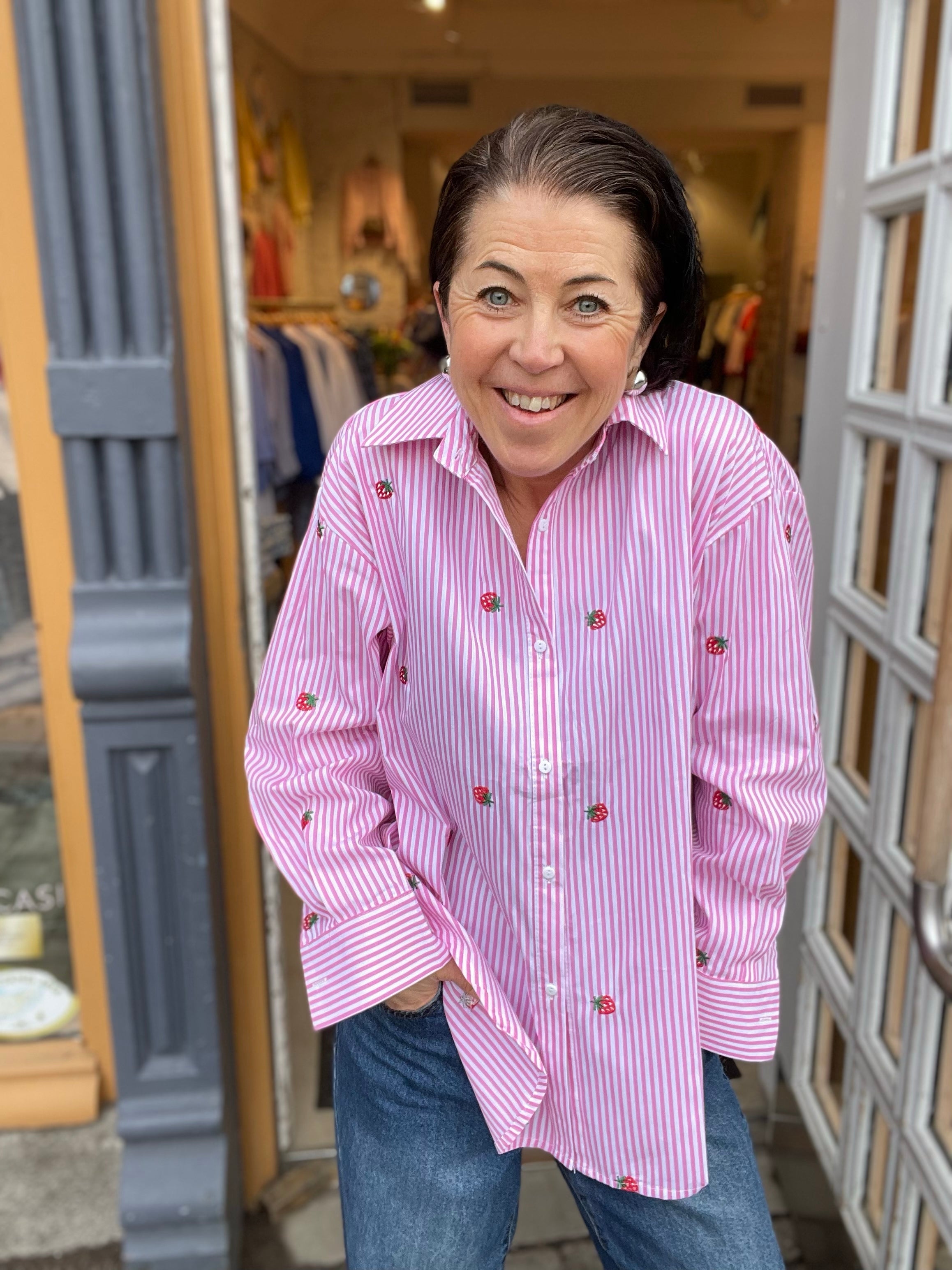 Leende kvinna framifrån i oversized rosa och vit randig skjorta med broderade jordgubbar, händerna i fickorna, stylade med blå jeans. 