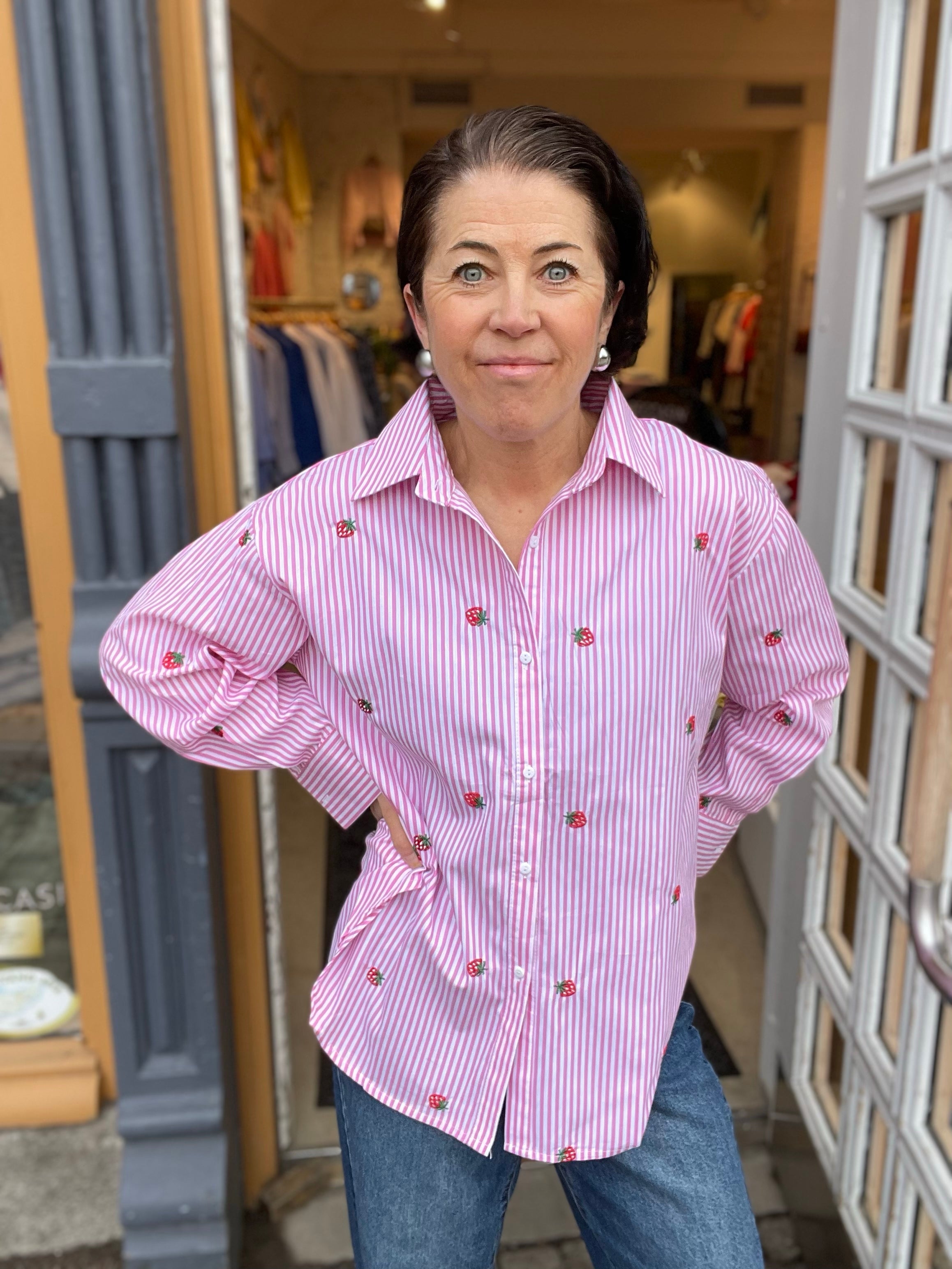 Kvinna med händerna i sidorna i rosa och vit randig skjorta med broderad jordgubbar, stylade med blå jeans, fotograferad framför butiksdörren utomhus.