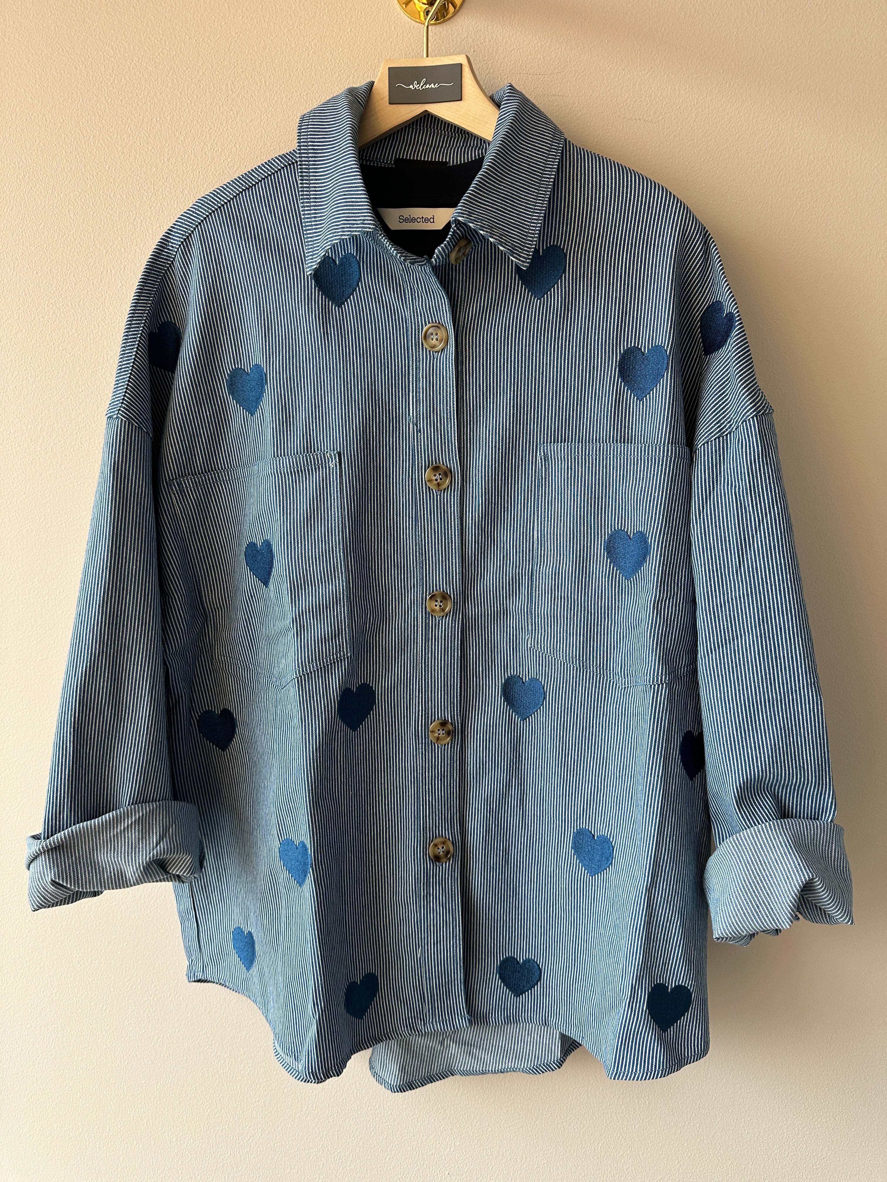 Blåvitrandig skjortjacka med hjärtmönster hängd på galge, helbild framifrån mot ljus vägg.