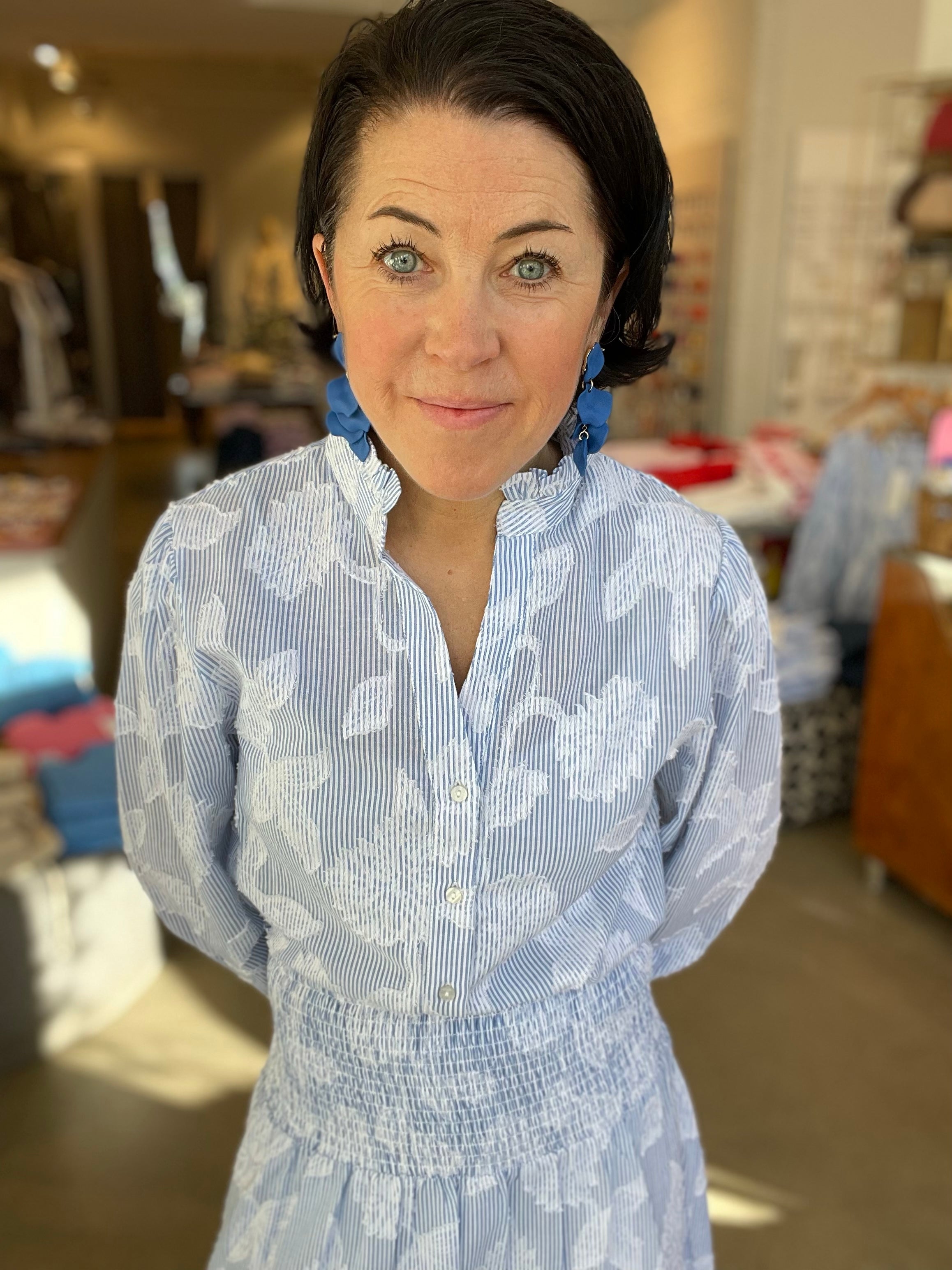  Kvinna framifrån med ett leende i blåvit randig blus med stora vita blomtryck, stående krage med v-ringning och puffiga ärmar, instoppad i matchande blåvit randig kjol med smockad midja, stylade med blå geometriska örhängen, inomhus i butik.