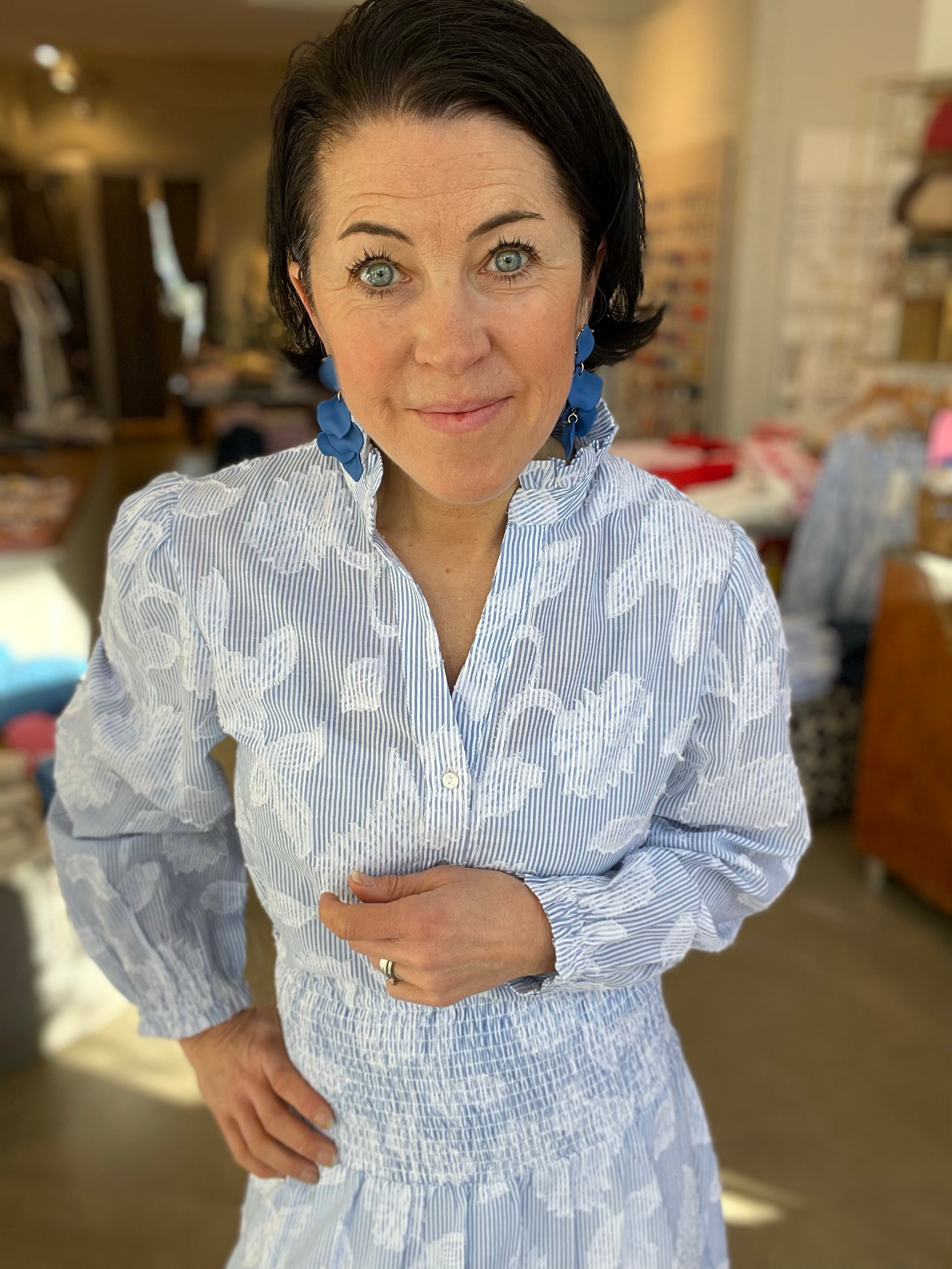 Kvinna närbild framifrån med handen vid midjan och ett leende i blåvit randig blus med stora vita blomtryck och stående krage, instoppad i matchande blåvit randig kjol med smockad midja, stylade med blå geometriska örhängen och ring, inomhus i butik.