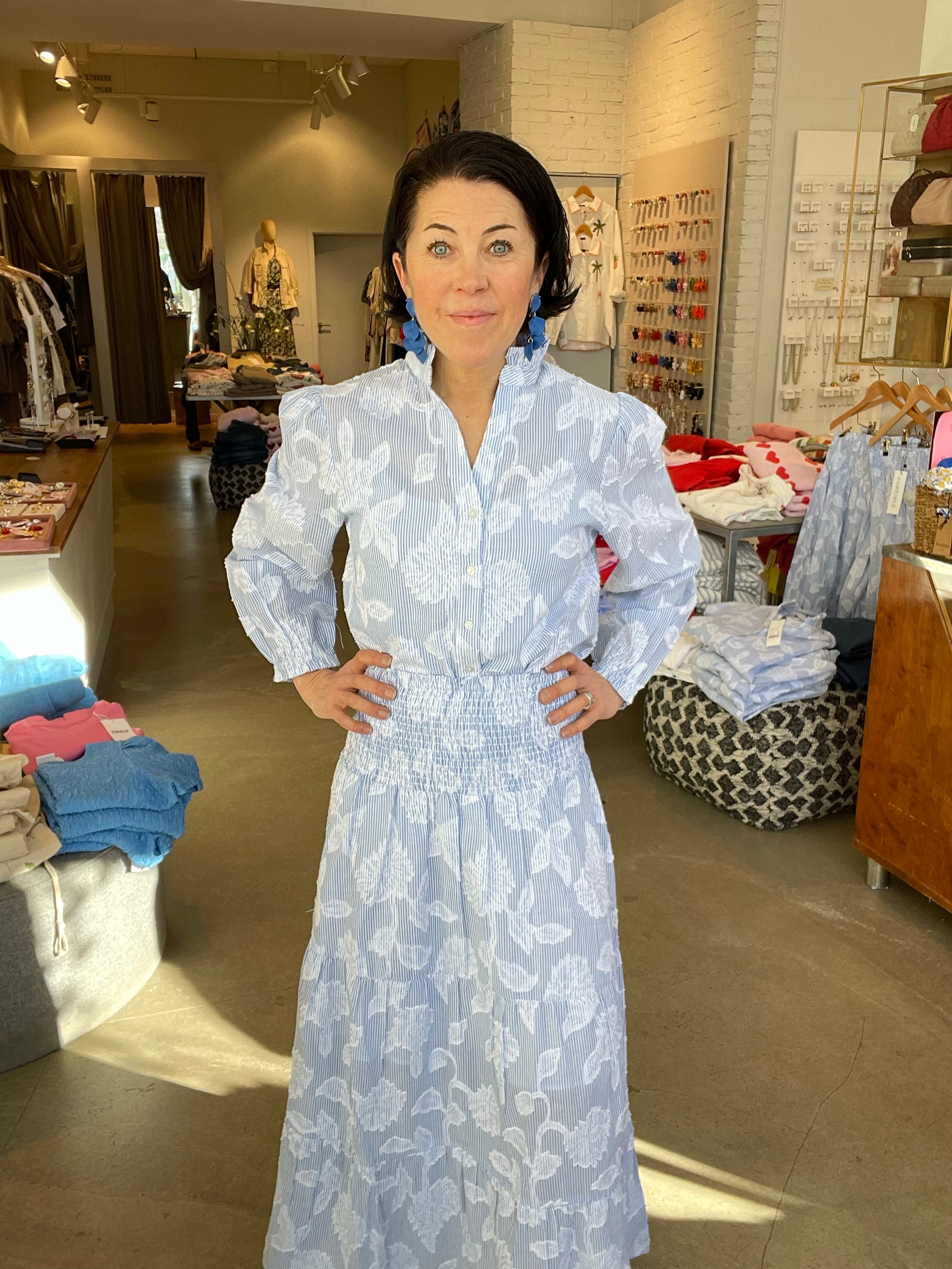 Kvinna framifrån med händerna i sidorna i blåvit randig blus med stora vita blomtryck och puffiga långa ärmar, instoppad i matchande blåvit randig kjol med smockad midja, stylade med blå geometriska örhängen, inomhus i butik med klädstativ i bakgrunden.