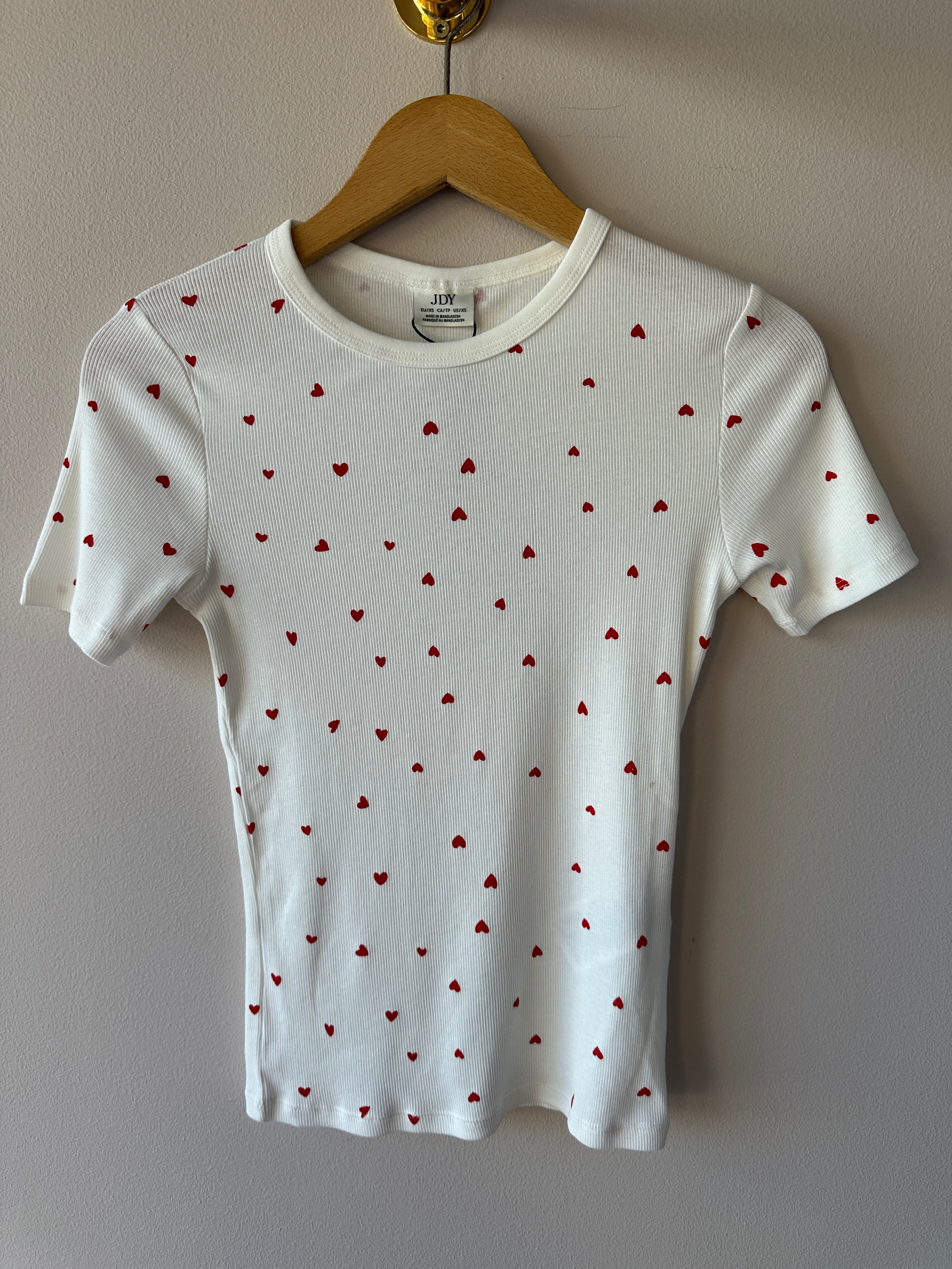 Vit ribbad t-shirt med små röda hjärtan över hela tyget, rund hals och korta ärmar hängande på galge mot vit vägg.