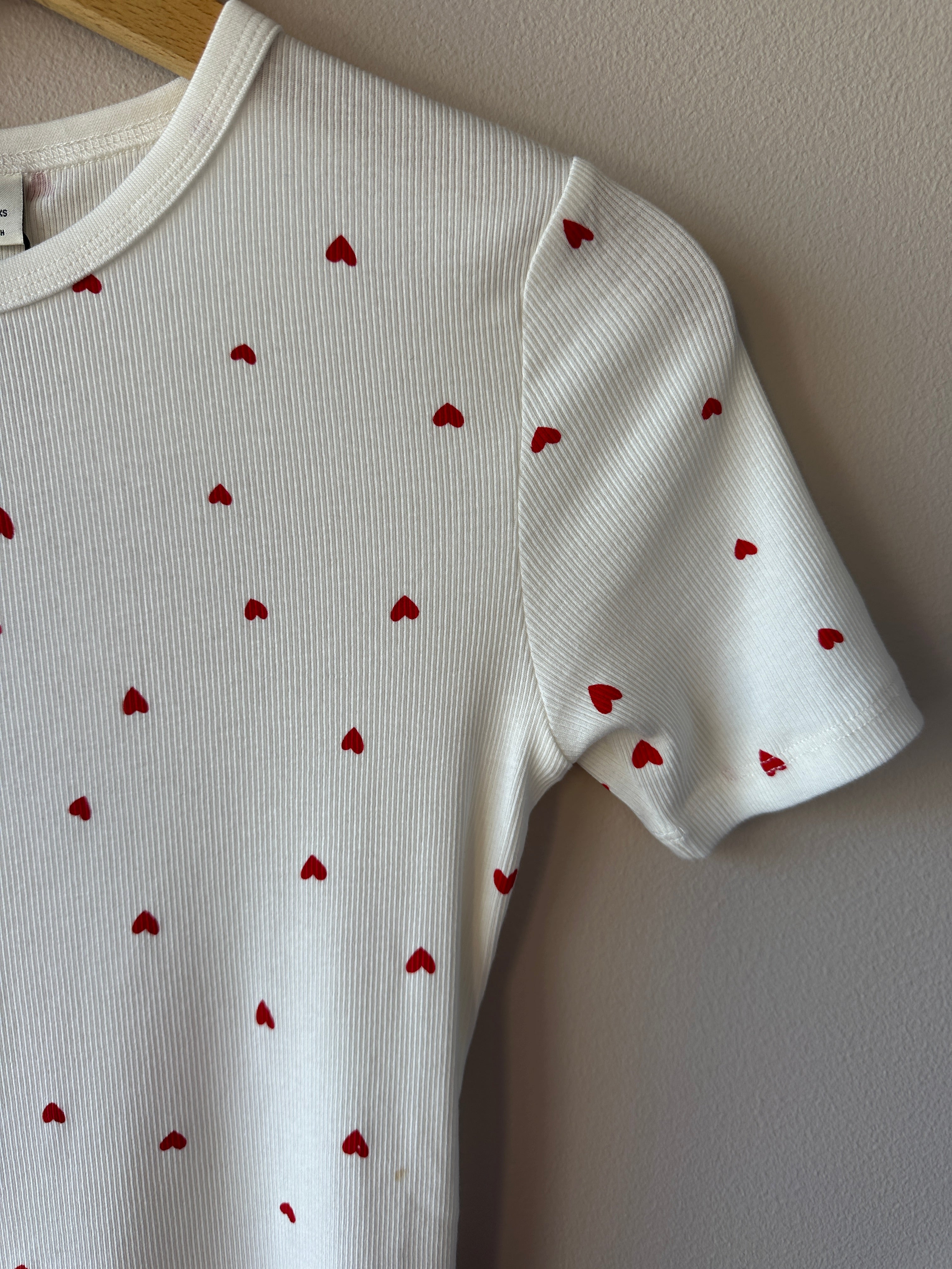 Närbild på vit ribbad t-shirt som visar små röda hjärtan över hela tyget och ribbad rund hals, hängande på galge.