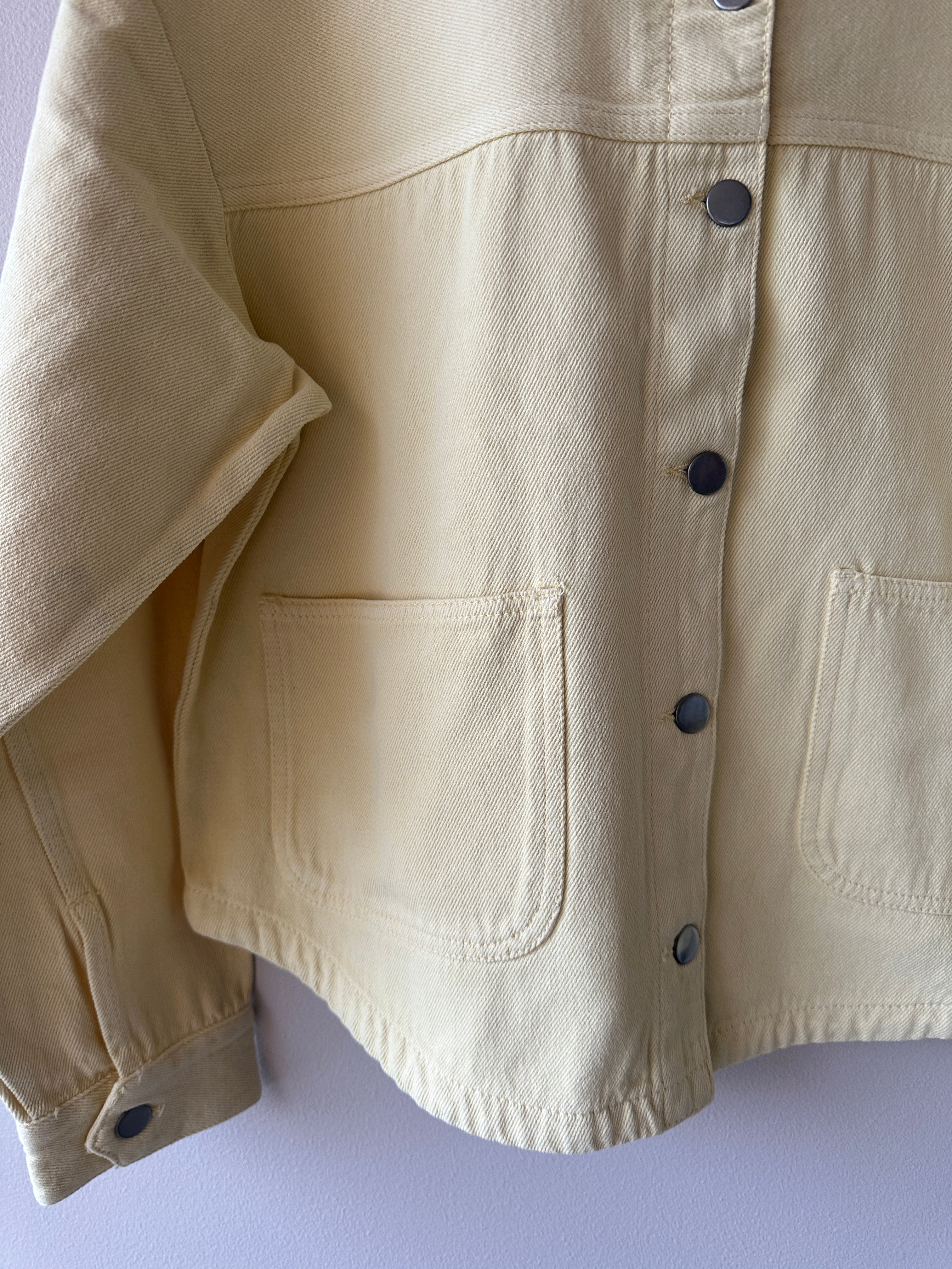 Närbild på framsidan av crèmefärgad shacket i denimtyg med mörka tryckknappar och stora patchfickor.