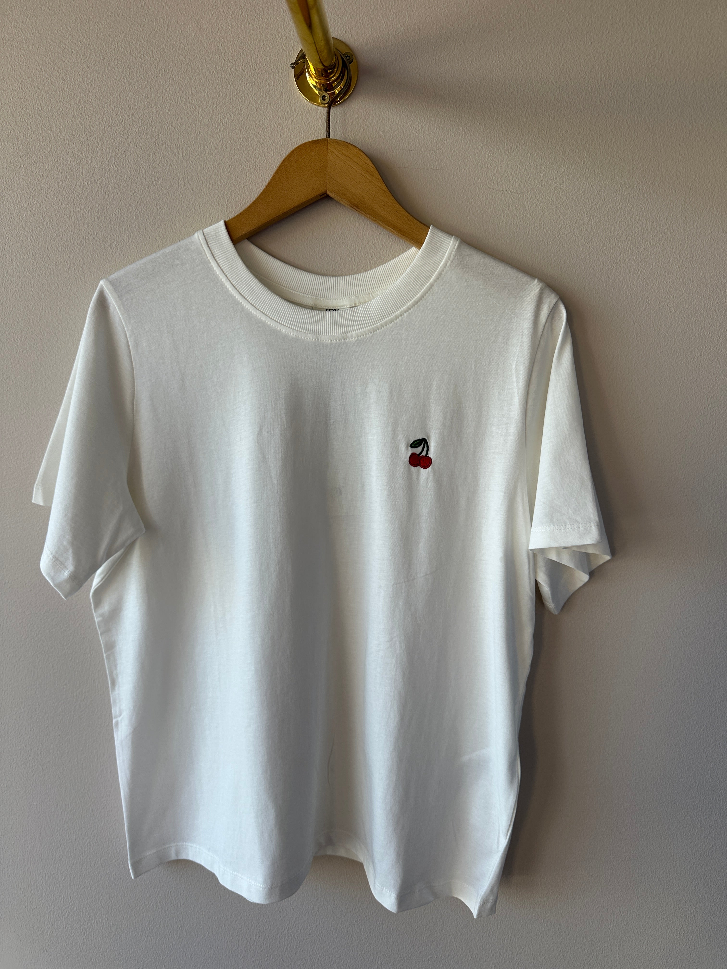 Vit oversized t-shirt med litet rött körsbroderi på bröstet, rund hals och korta ärmar hängande på galge mot vit vägg.