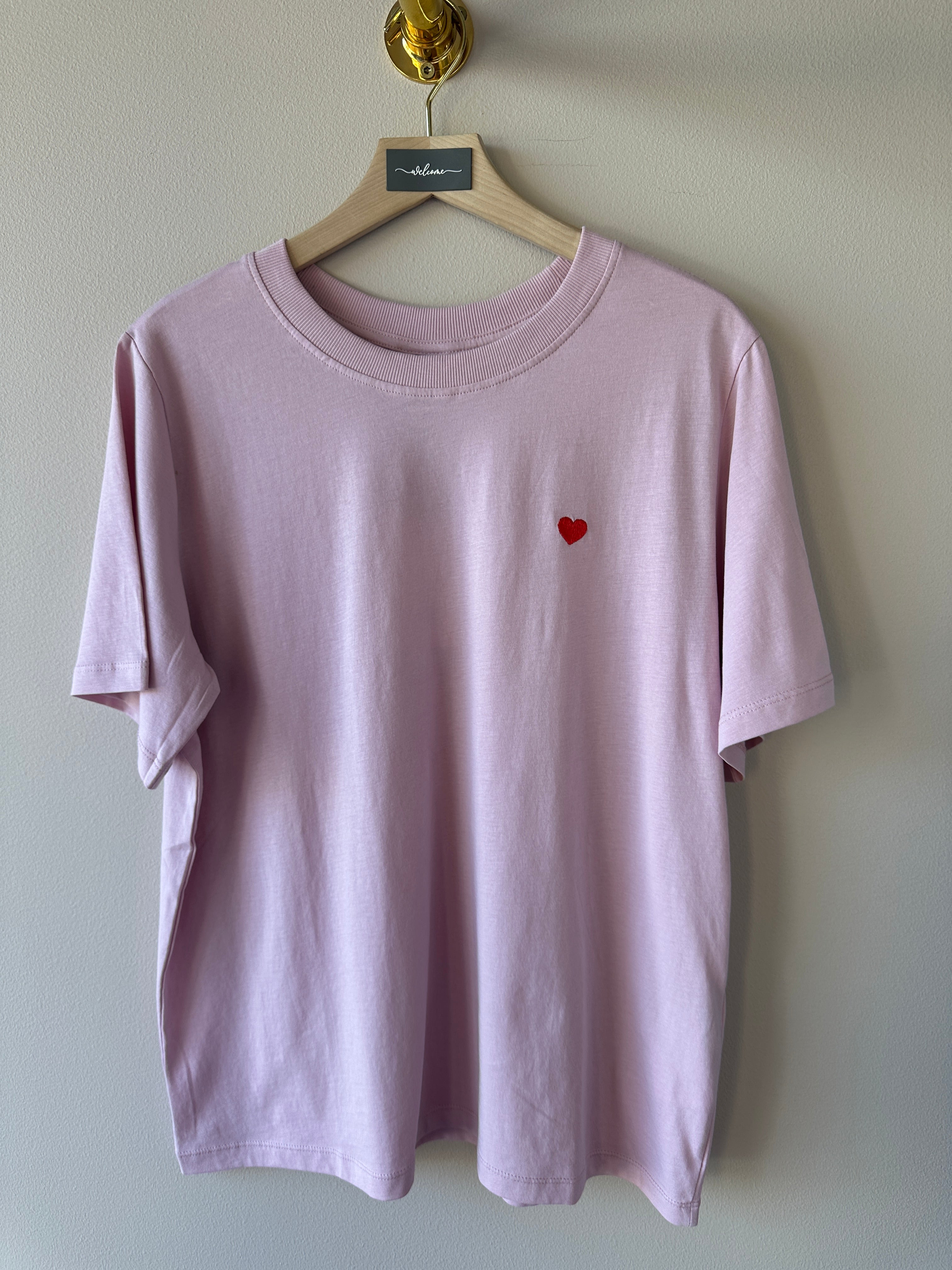 Ljuslila oversized t-shirt med litet rött hjärtbroderi på bröstet, rund ribbad hals och korta ärmar hängande på galge mot vit vägg.