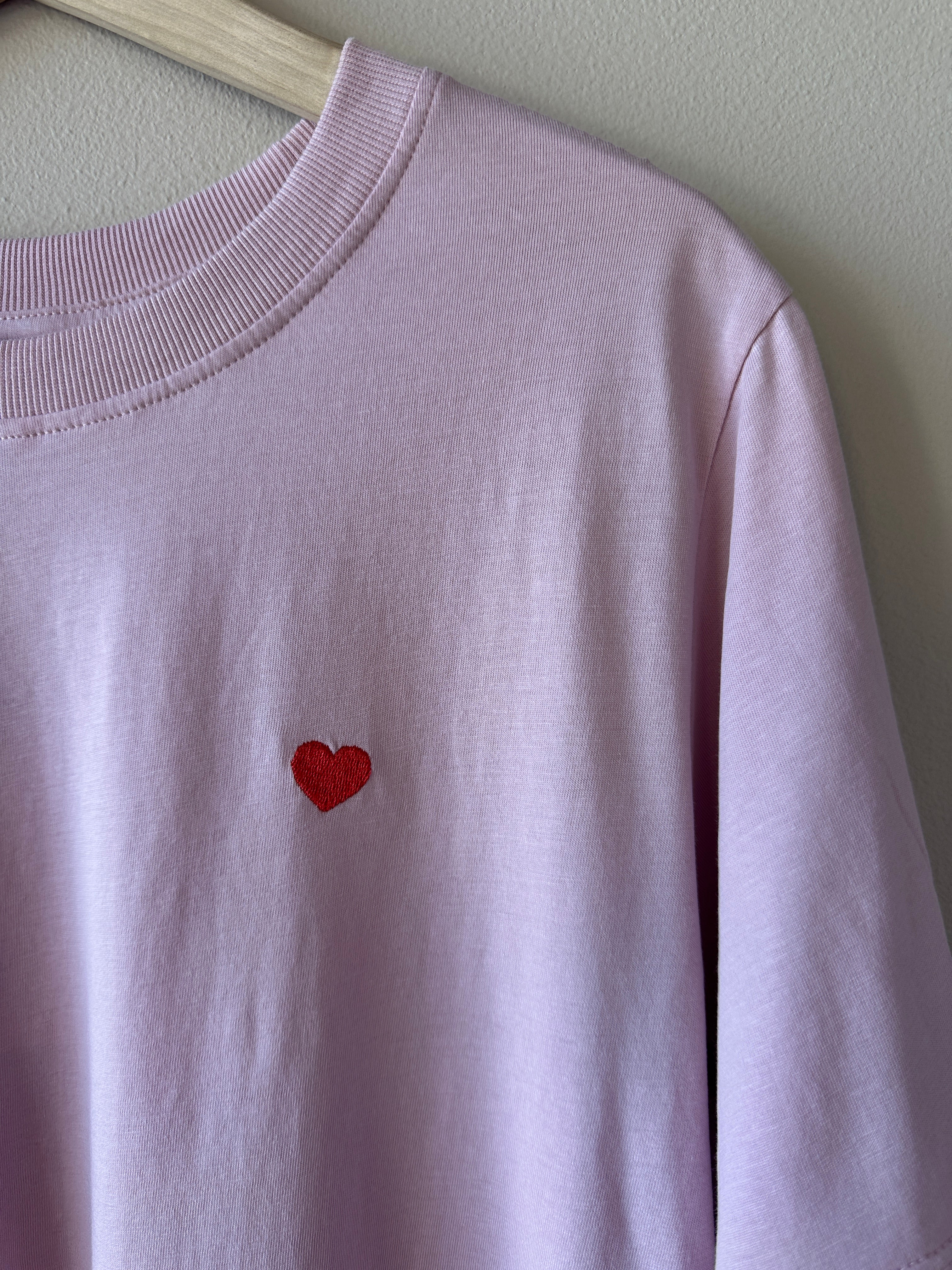 Närbild på bröstet av ljuslila t-shirt som visar litet rött hjärtbroderi på mjukt lila bomullstyg med ribbad rund hals.