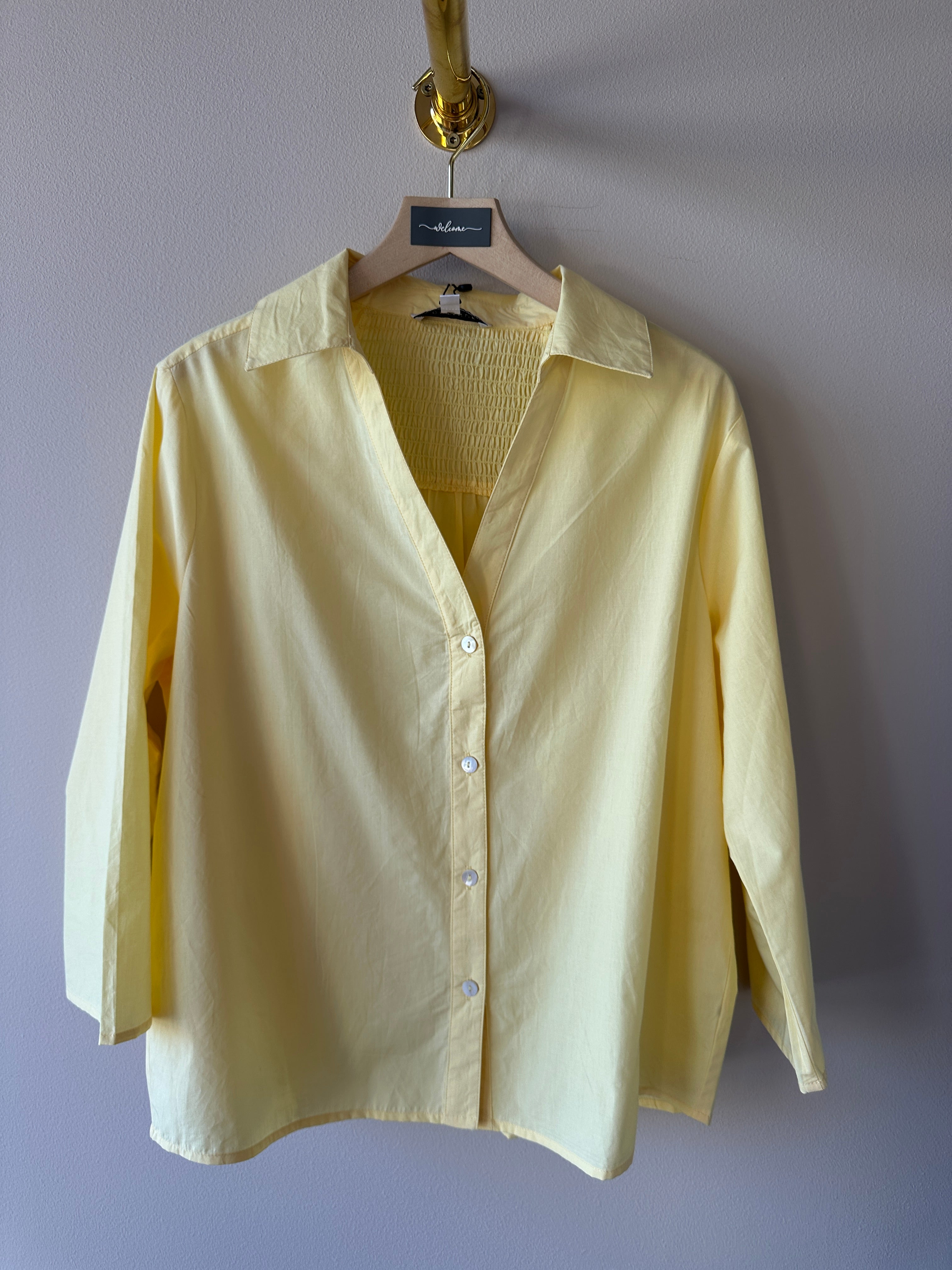 Gul oversized skjorta med klassisk krage, djup v-ringning och knappar fram hängande på galge mot grå vägg.