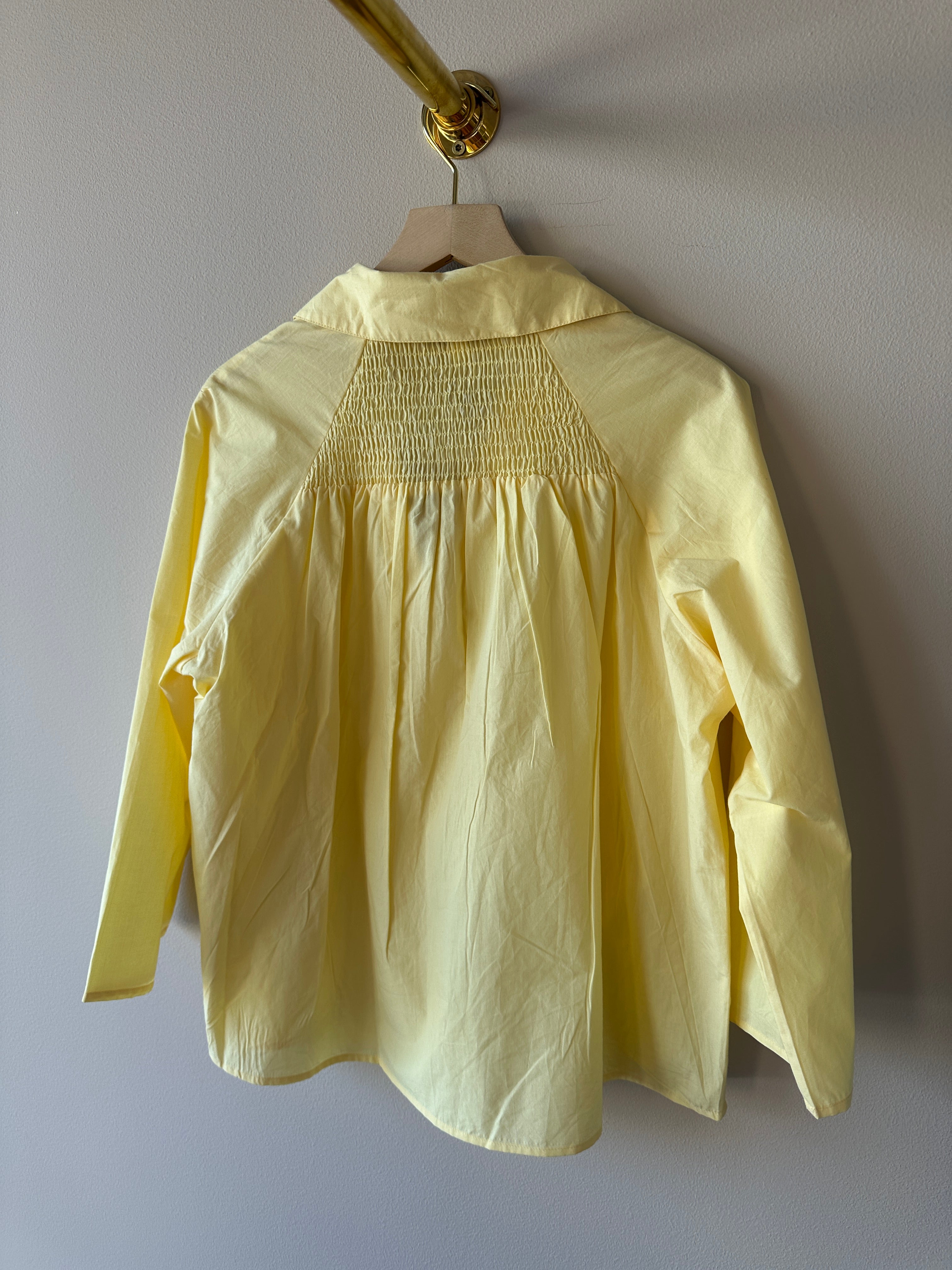 Baksida av gul oversized skjorta med stor smockad panel över övre ryggen och vida långa ärmar, hängande på galge mot grå vägg.