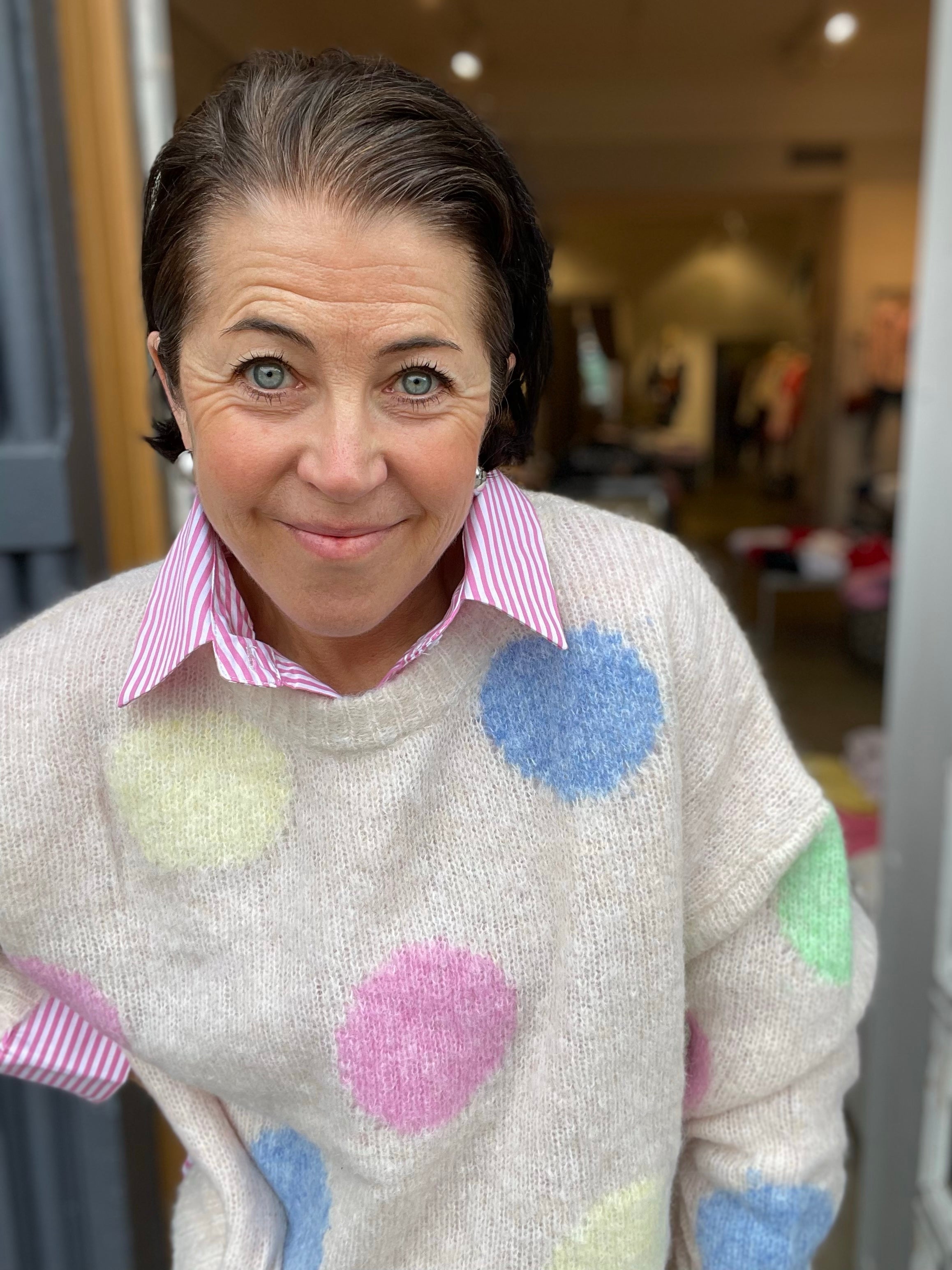 Kvinna framifrån i oversized ecru stickad tröja med stora pastellfärgade prickar och randig rosa skjorta under, händerna i sidorna, fotograferad i butiksdörren.
