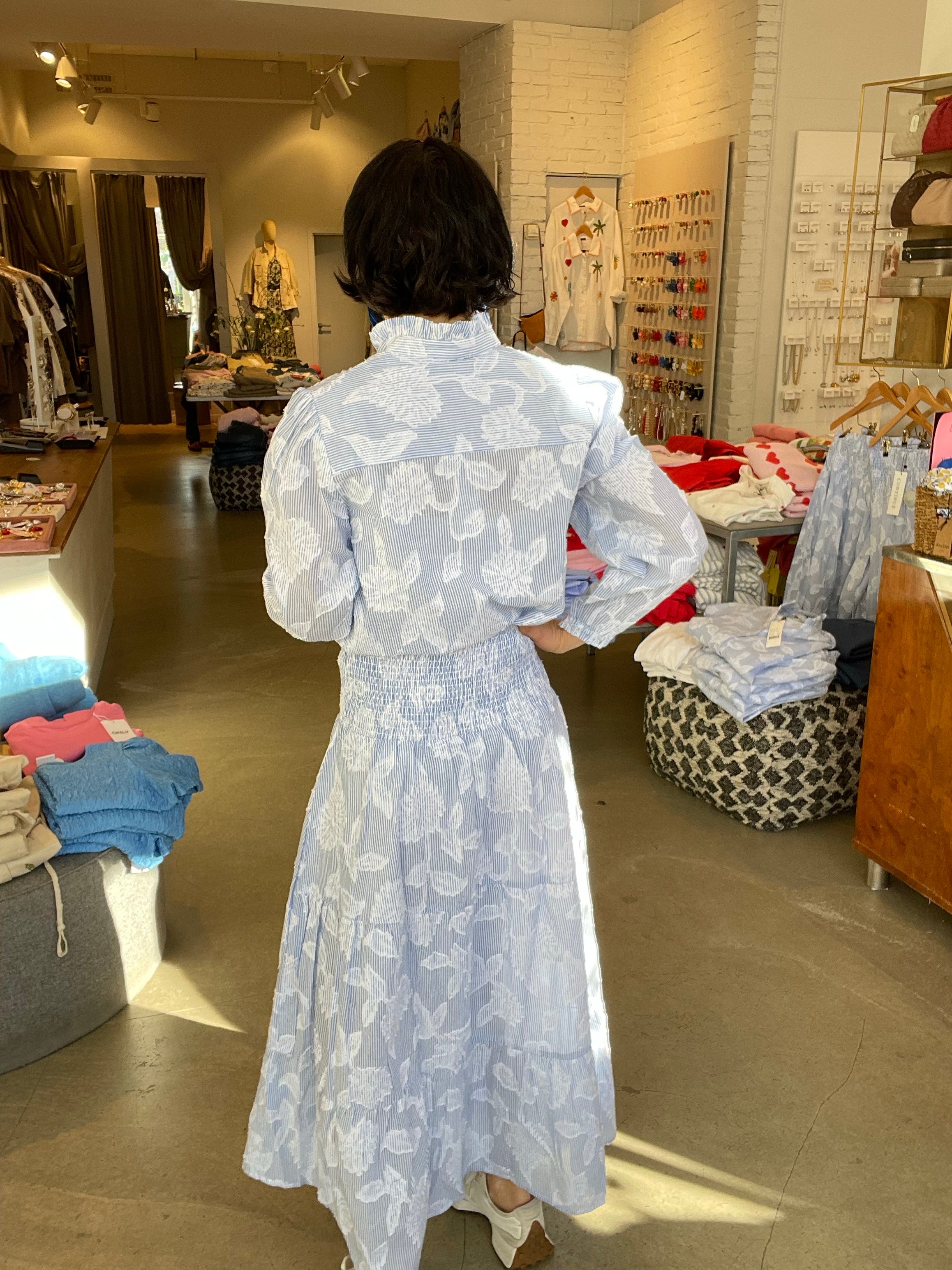 Kvinna bakifrån med handen i sidan i blåvit randig midi-kjol med stora vita blomtryck, smockad midja och vid kjolkant med volangkant, stylade med matchande blåvit randig blus och vita skor, inomhus i butik med klädstativ i bakgrunden.