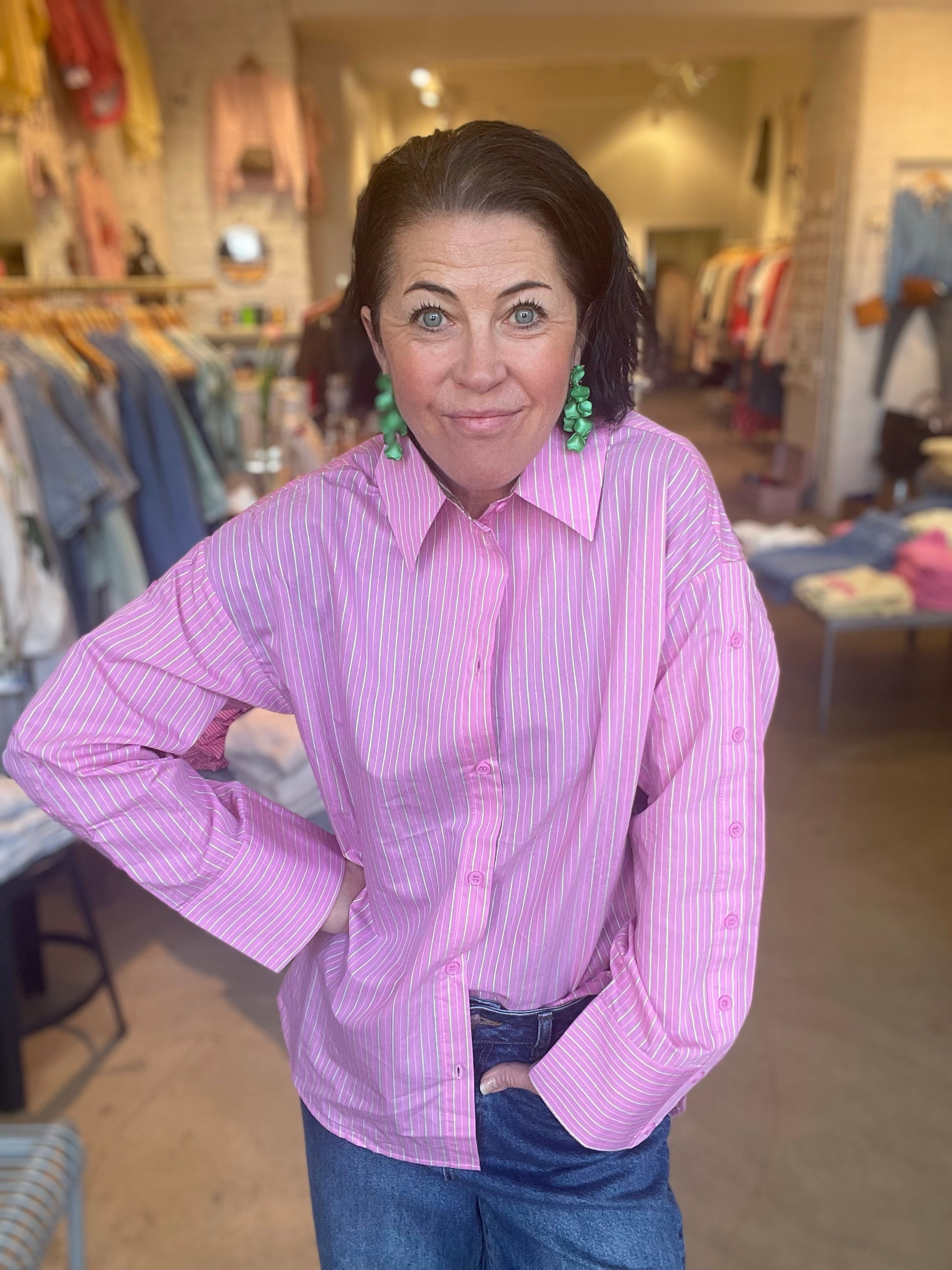 Kvinna med handen i fickan i oversized cerise och vit randig skjorta med klassisk krage och långa ärmar, stylade med mörka jeans och gröna örhängen,  fotograferad i butiksmiljö.