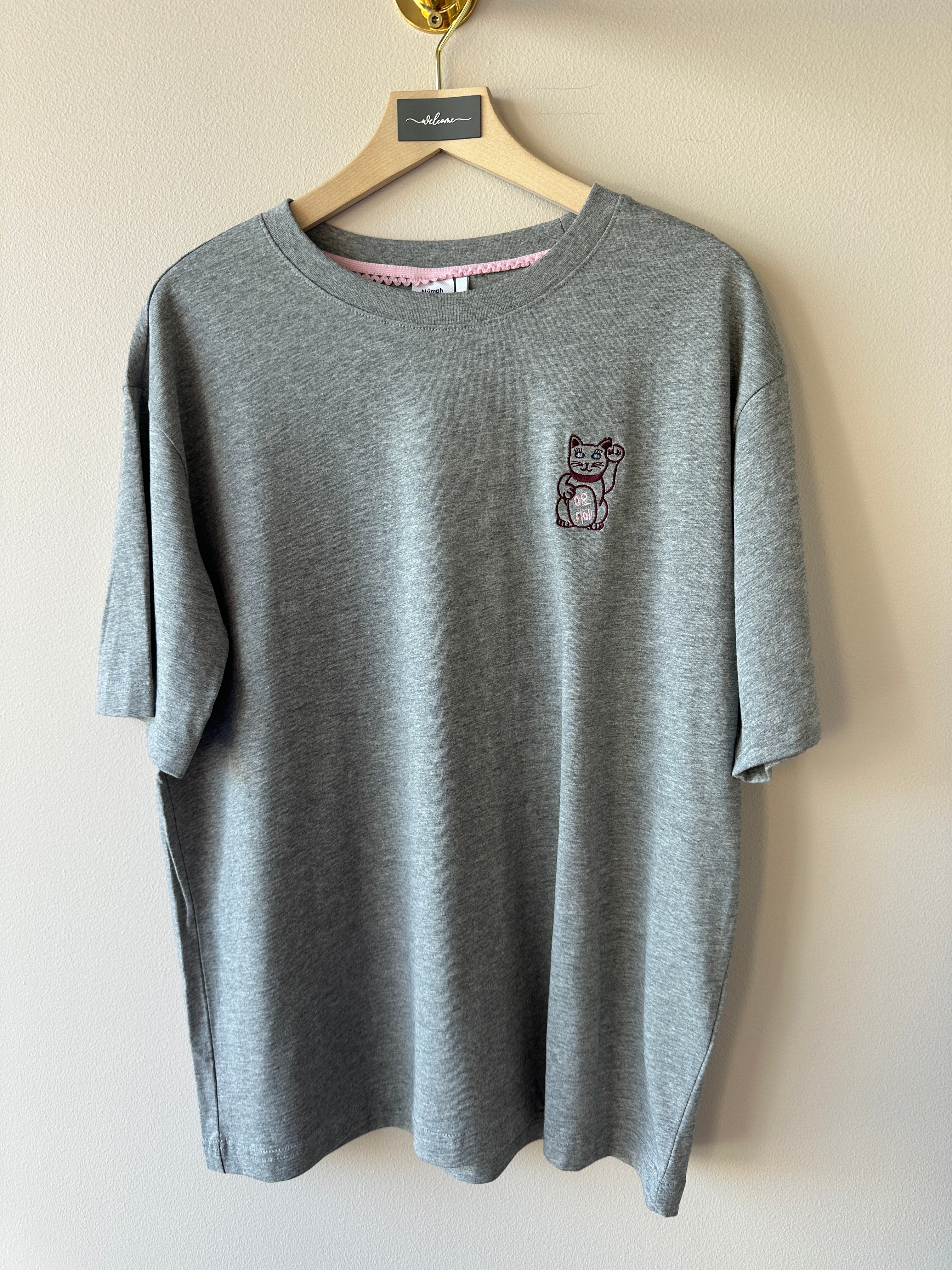 Gråmelerad oversized t-shirt med litet broderi av en japansk lyckokatt på bröstet, rund hals och korta ärmar hängande på galge mot beige vägg.