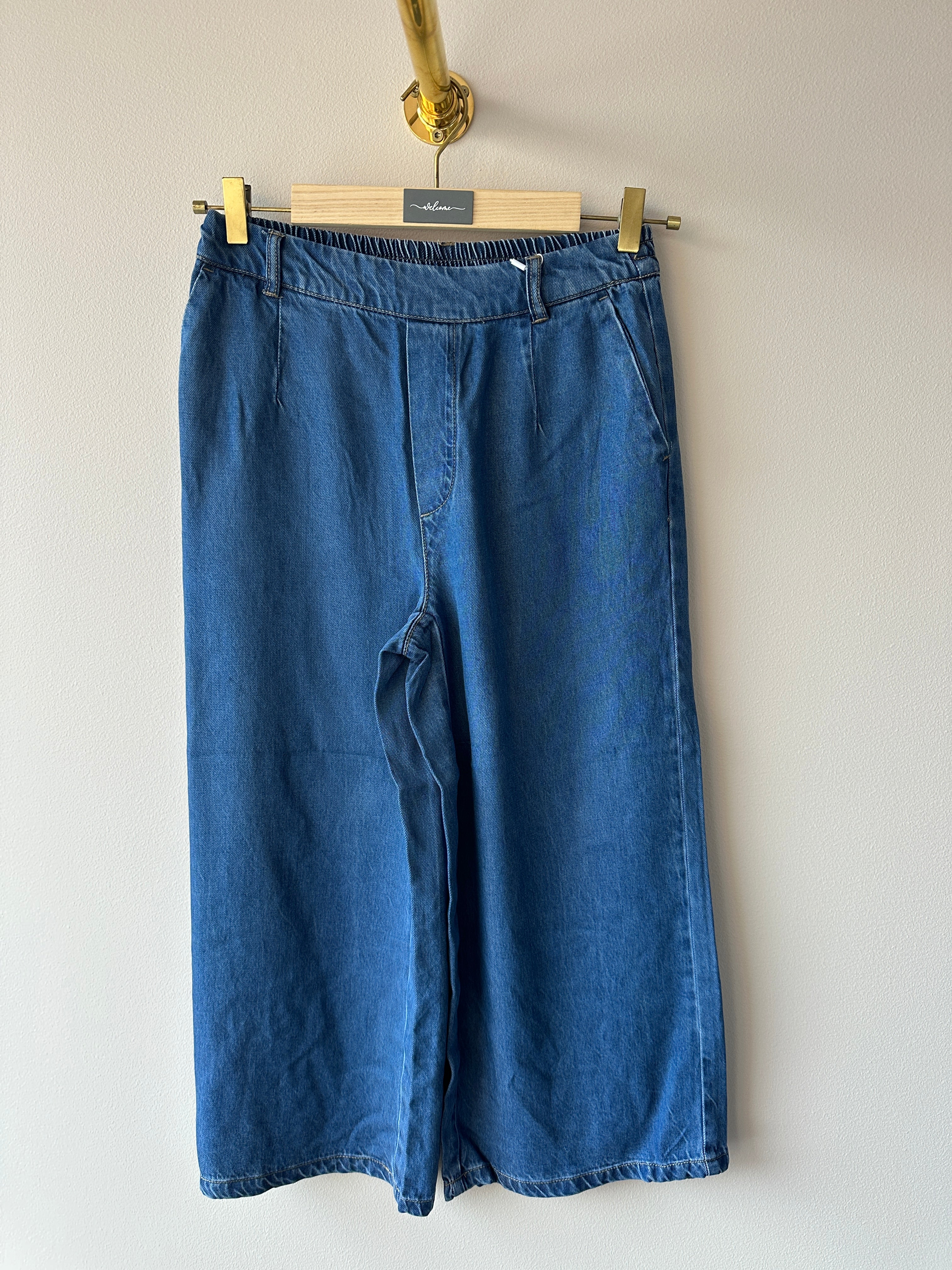 Mellanblå denimculottes med elastisk midja, plisserade veck fram och vida ben, hängande på galge mot vit vägg, sedda framifrån.