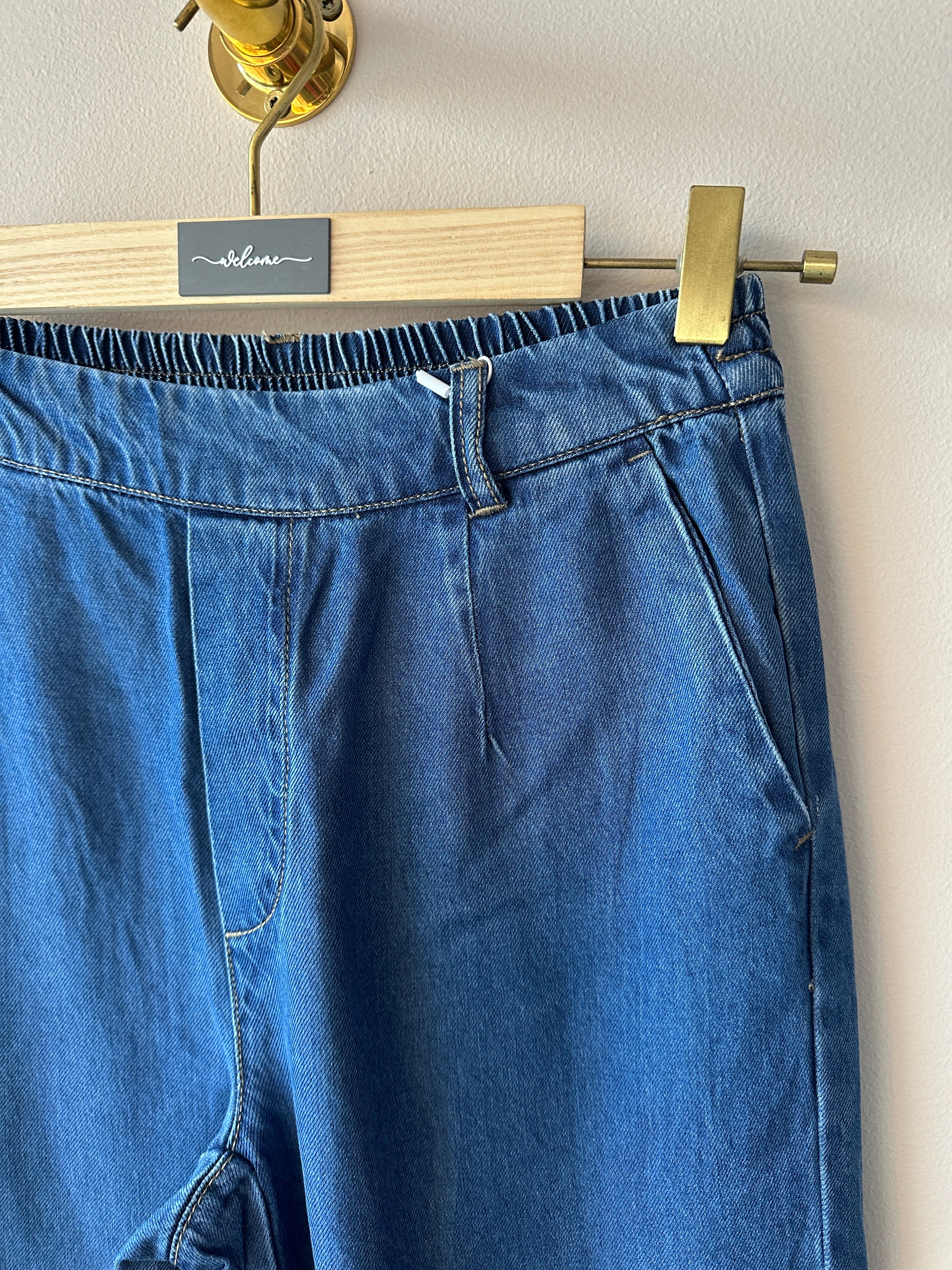 Närbild på midja och övre del av mellanblå denimculottes som visar elastisk midja med synliga stygn, plisserade veck fram och sneda sidofickor.