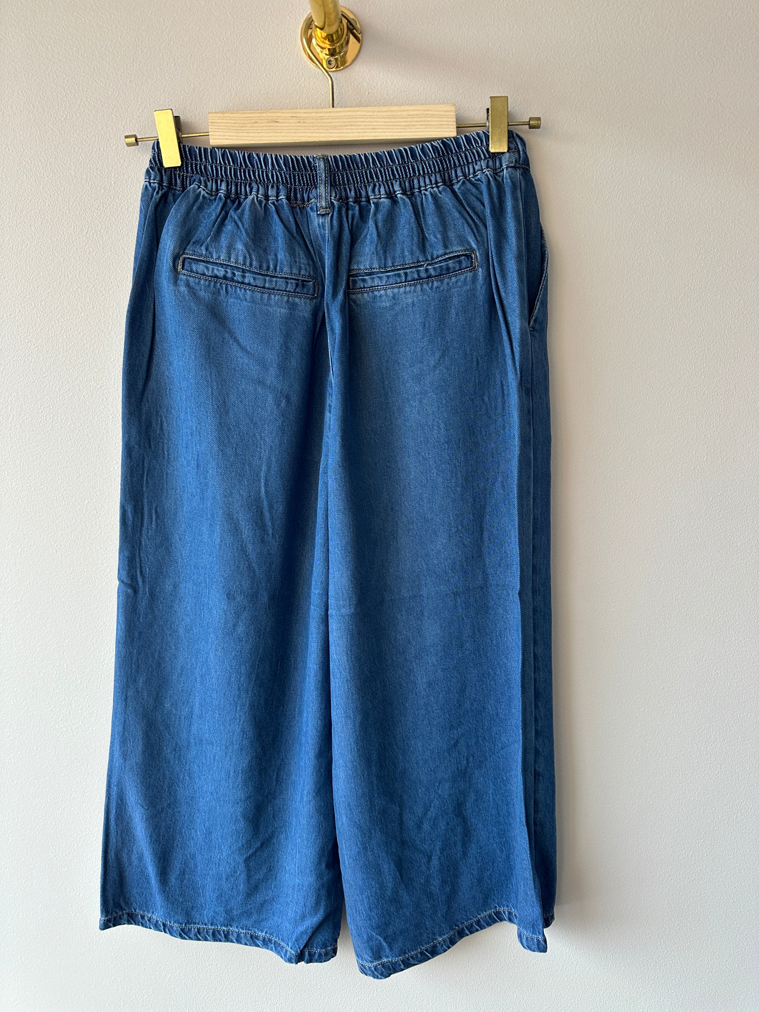 Baksida av mellanblå denimculottes med elastisk midja och bakficka med klaff, vida ben med rak avslutning vid vaden, hängande på galge mot vit vägg.