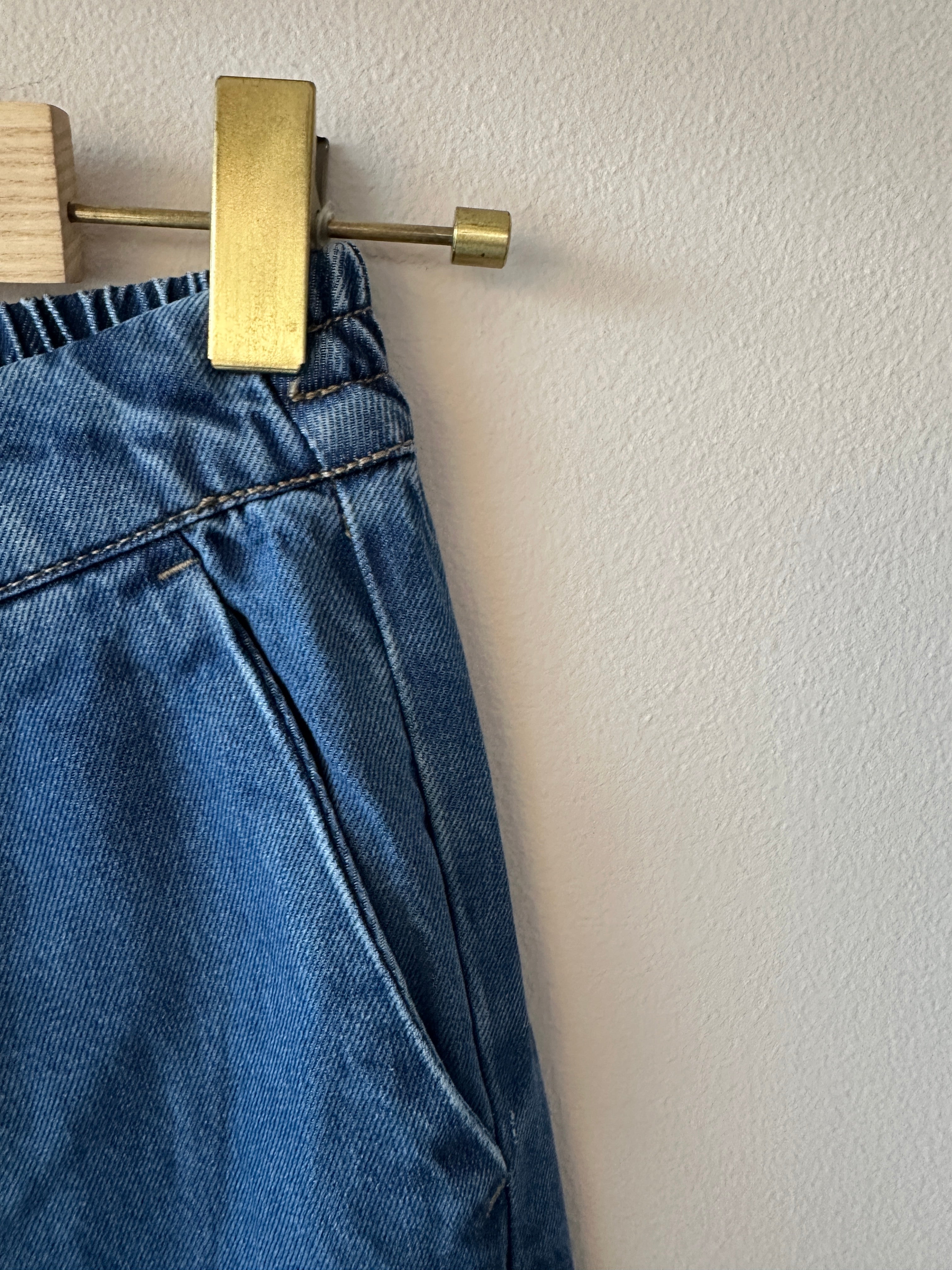 Närbild på sidan av mellanblå denimculottes som visar elastisk midja med synliga stygn och plisserat veck fram i mellanblå denim.