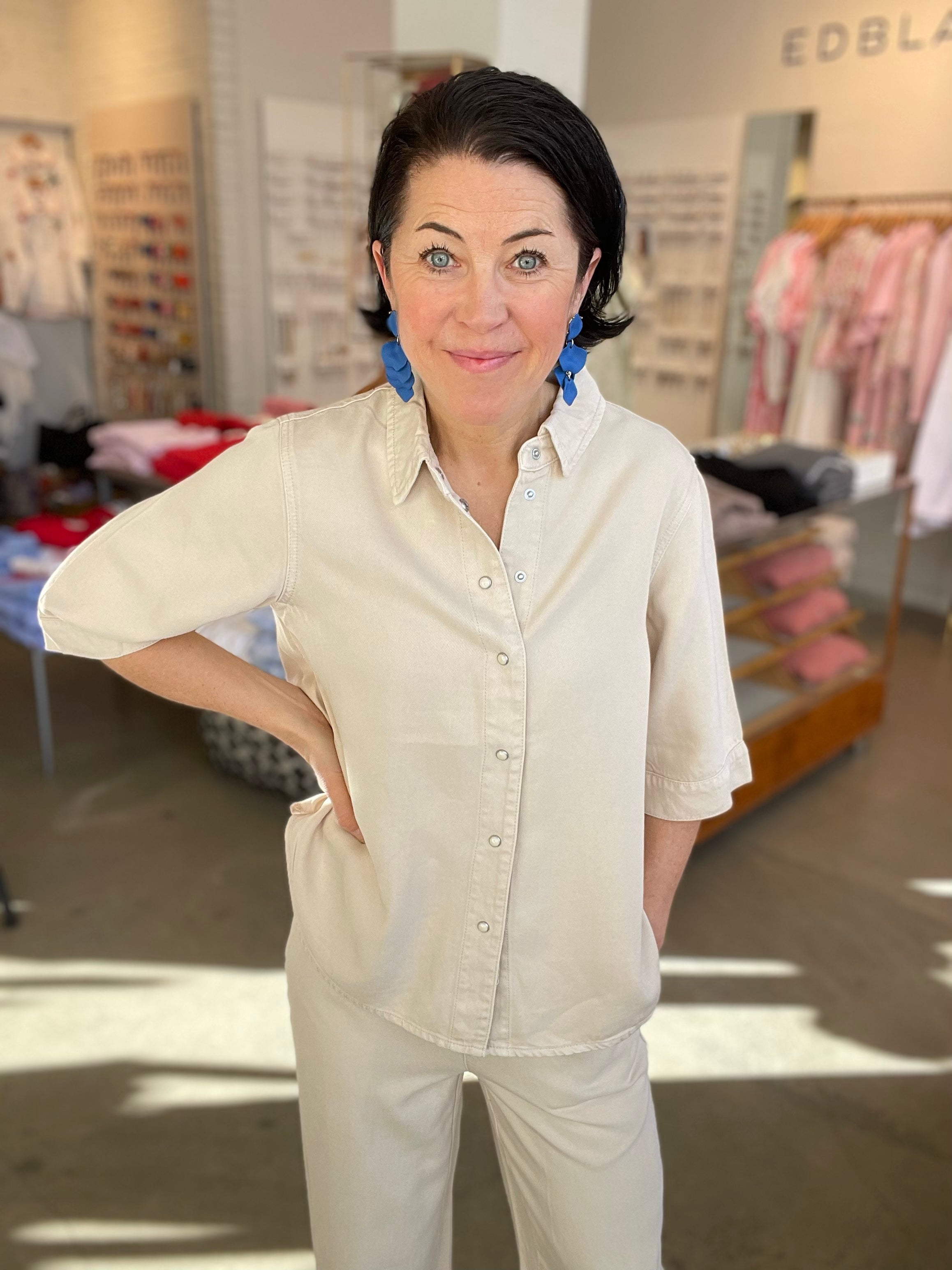 Kvinna framifrån med handen i sidan i beige denimtopp med klassisk krage och tryckknappar i silver, stylade med beige jeans och blå blommörhängen, inomhus i butik.