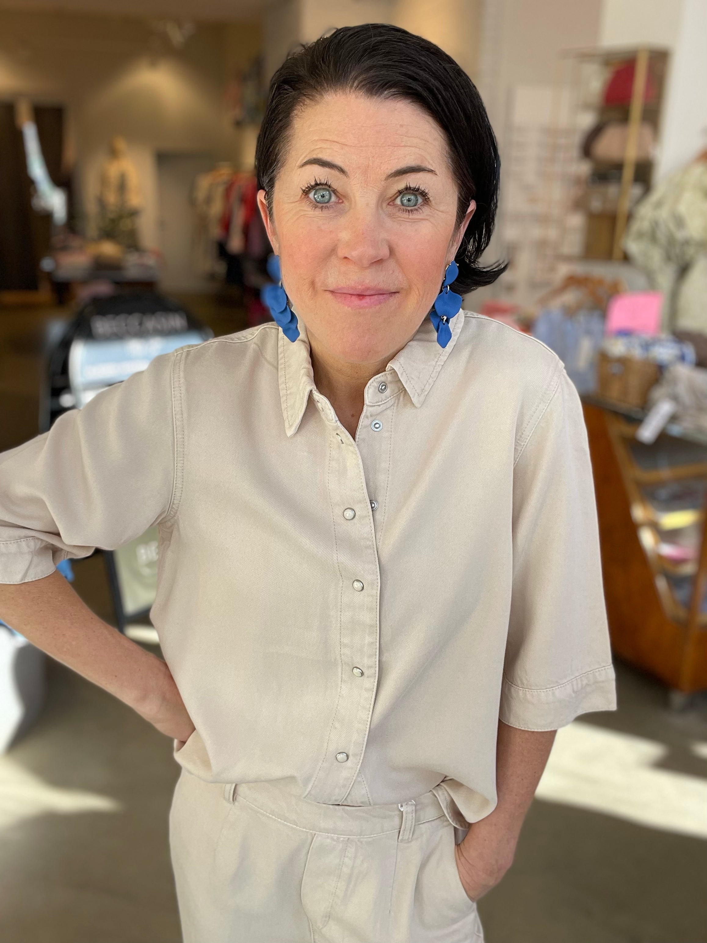 Kvinna närbild framifrån med handen i fickan i beige denimtopp med klassisk krage och tryckknappar i silver, stylade med beige jeans och blå blommörhängen, inomhus i butik.