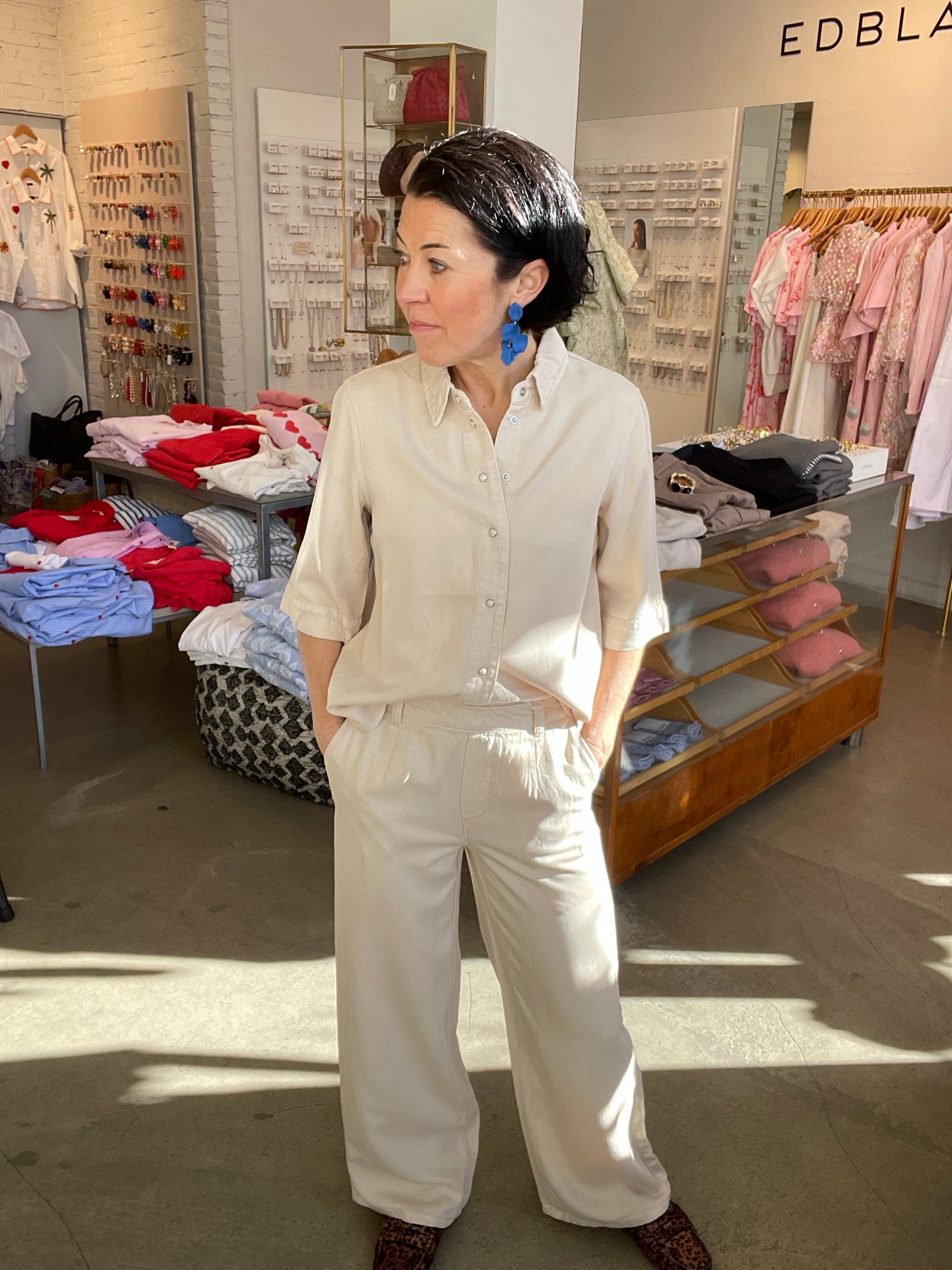 Kvinna i halvprofil med blicken åt sidan i beige wide leg-byxor med hög midja, stylade med matchande beige denimtopp, blå blommörhängen och leopardmönstrade skor, inomhus i butik.