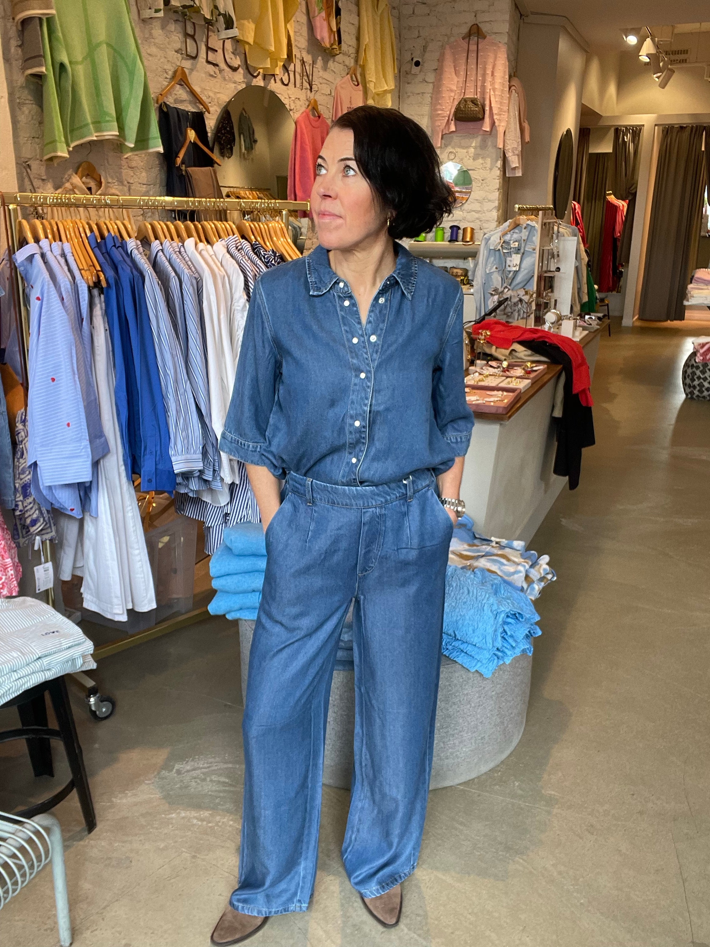Kvinna framifrån i vida mellanblå denimbyxor med hög midja och stora fickor, stylade med matchande denimskjorta och bruna boots, fotograferad i butiksmiljö med klädstativ i bakgrunden.