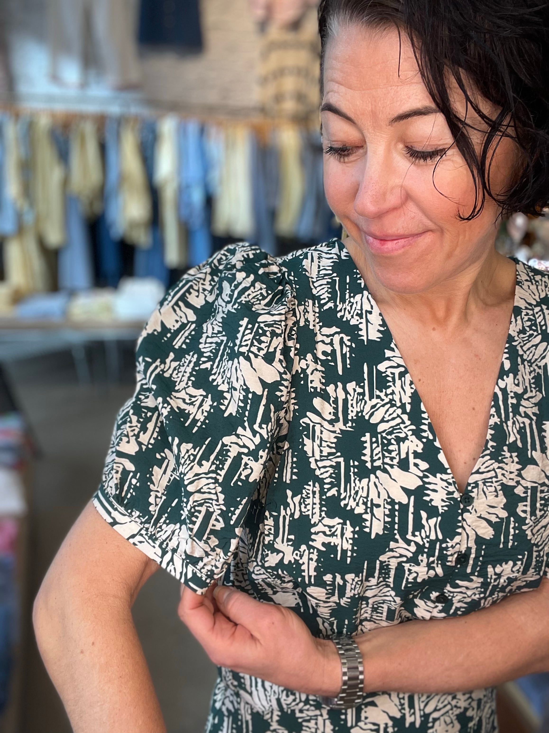 Kvinna som tittar ned i mörkgrön abstrakt topp med V-ringning och puffiga korta ärmar, närbild i butiksmiljö.