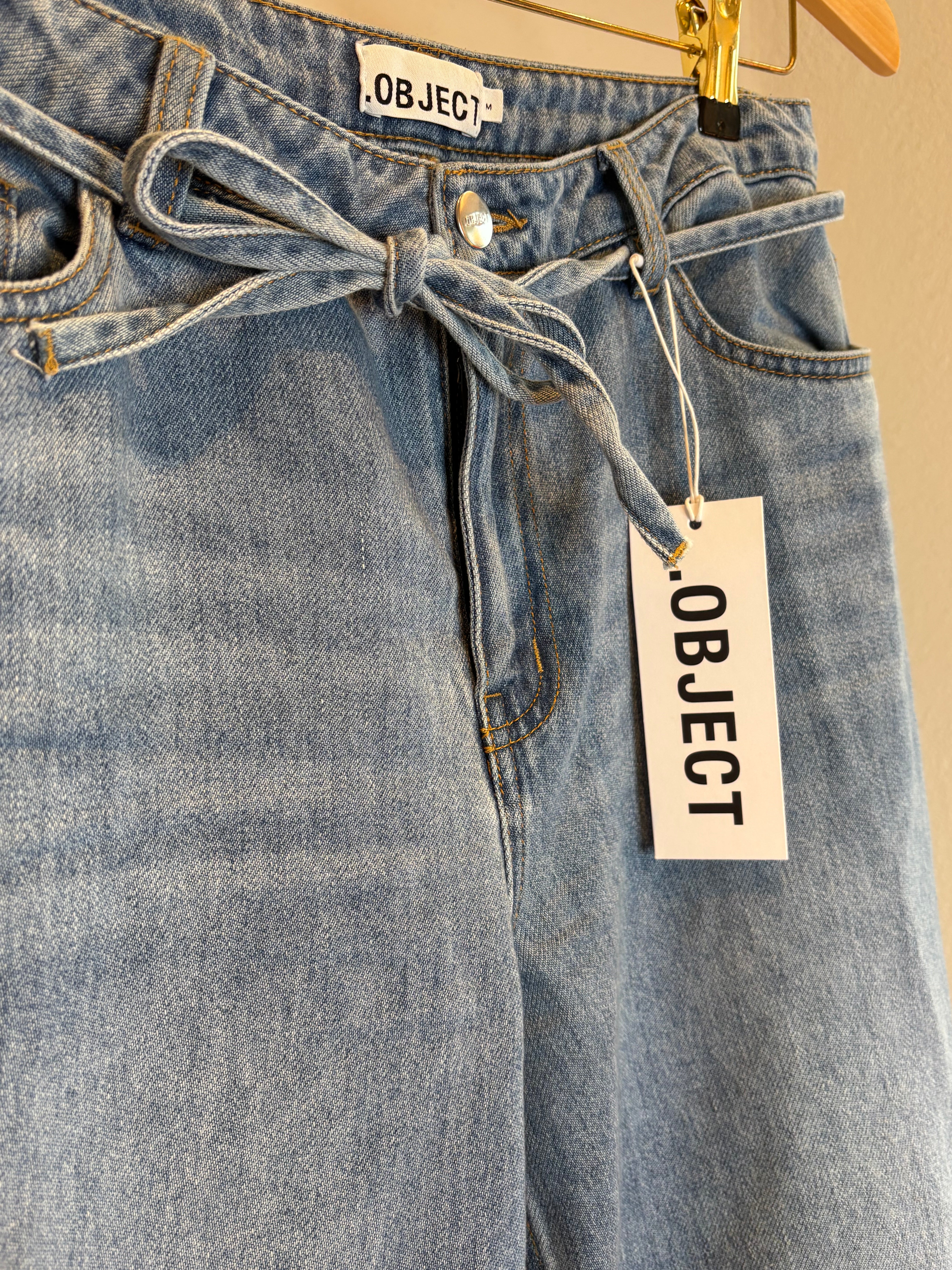 Objrosie Wide Jeans Light Blue Denim. Blåa jeans med snörning hängandes på en galge.