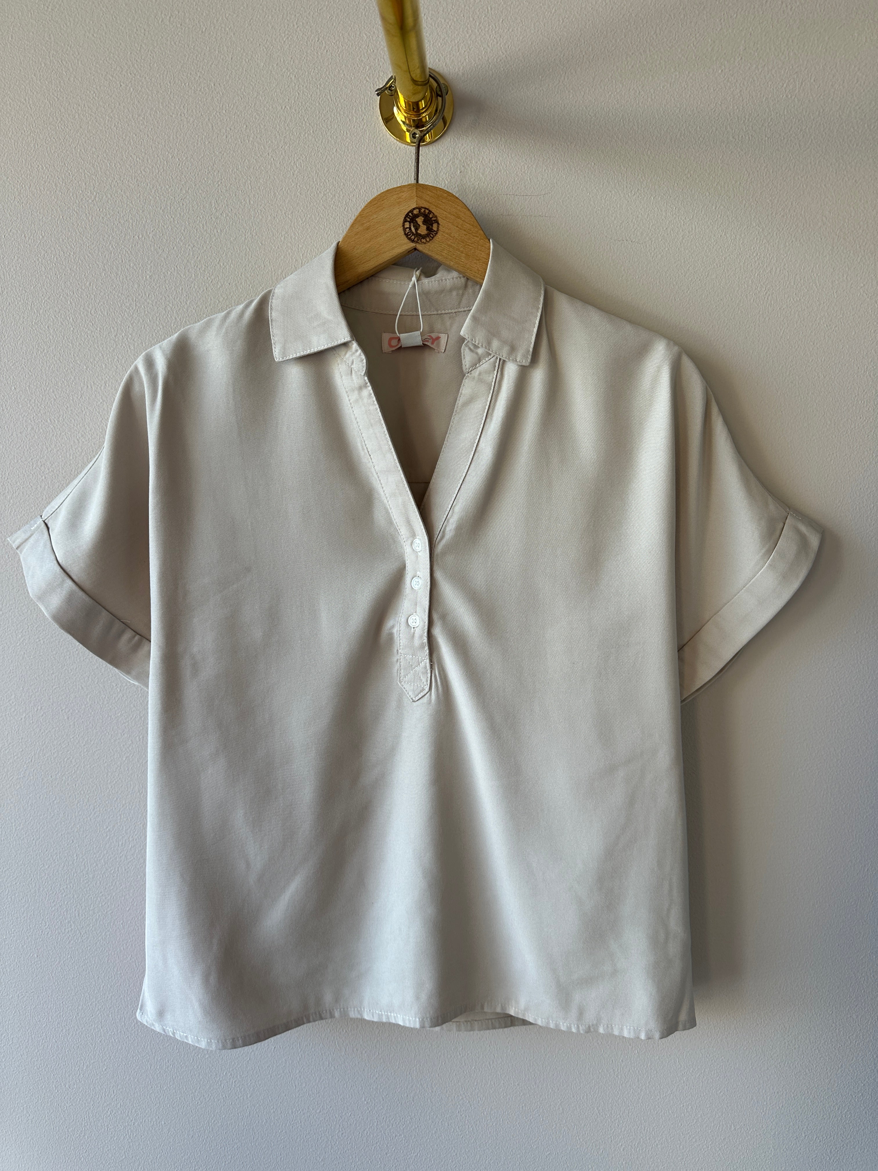 Beige kortärmad skjorta med djup v-ringning, knappar fram och lätt oversized passform hängande på galge mot vit vägg.