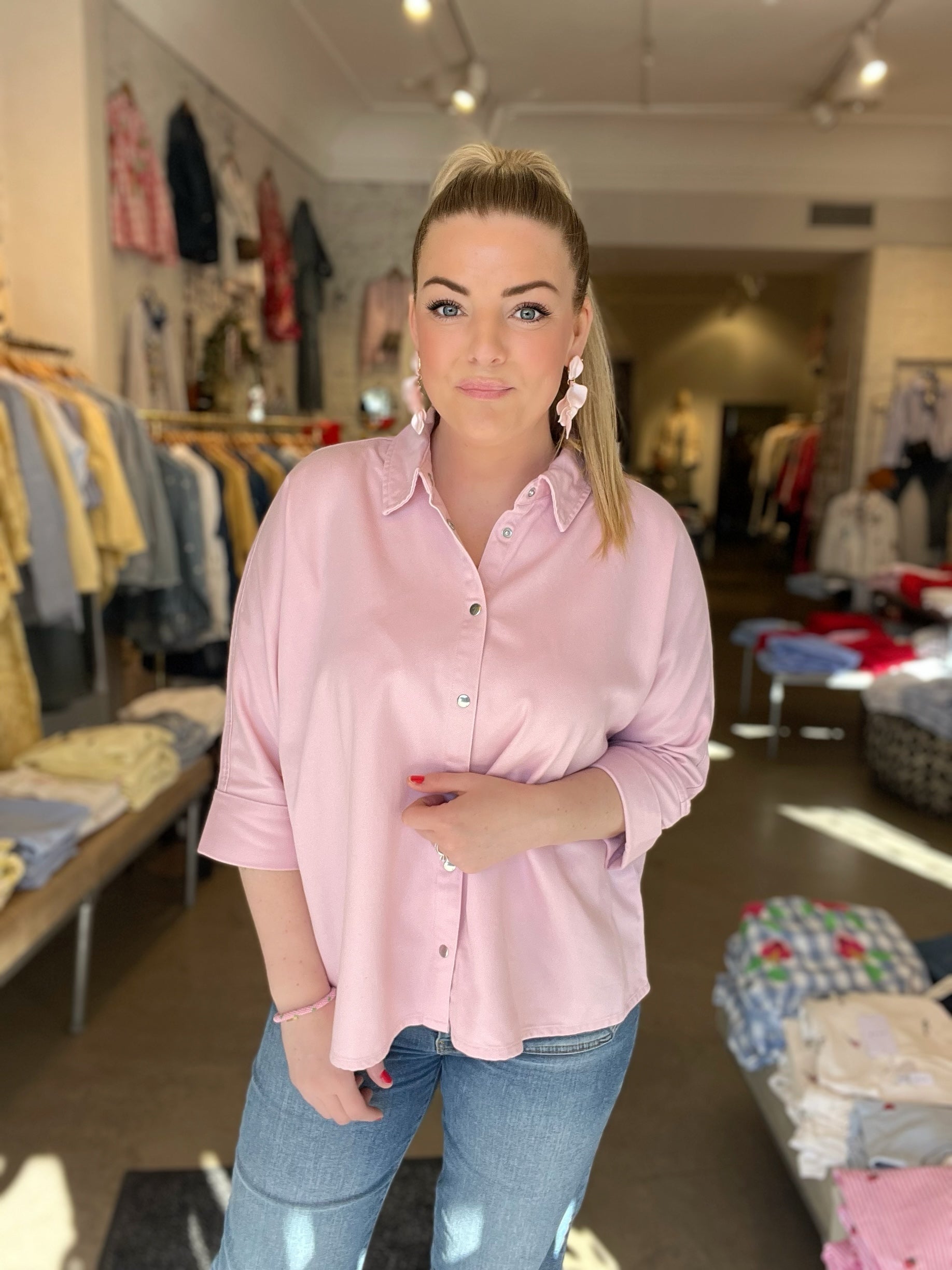 Kvinna framifrån i oversized rosa skjorta med klassisk krage och tryckknappar, stylade med blå jeans och rosa blommörhängen, inomhus med klädstativ i bakgrunden.