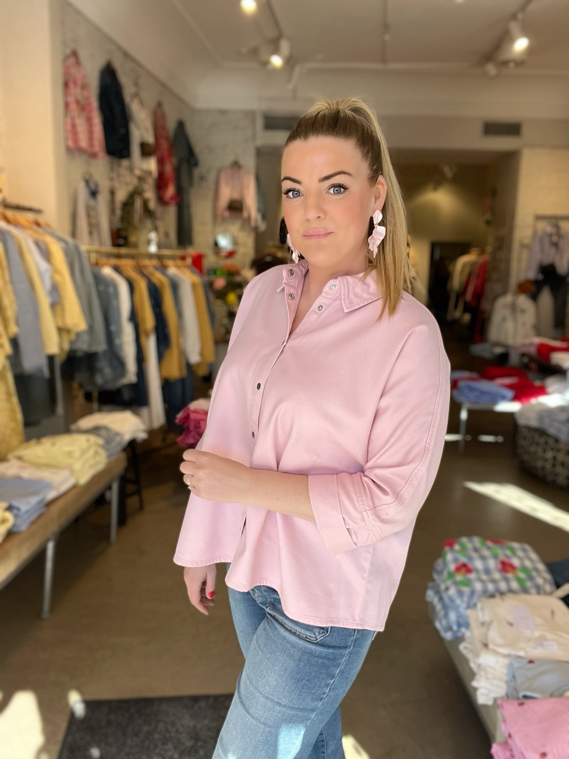 Kvinna i halvprofil i oversized rosa skjorta med 3/4-långa ärmar och uppvikta manschetter, stylade med blå jeans och rosa blommörhängen, inomhus med klädstativ i bakgrunden.