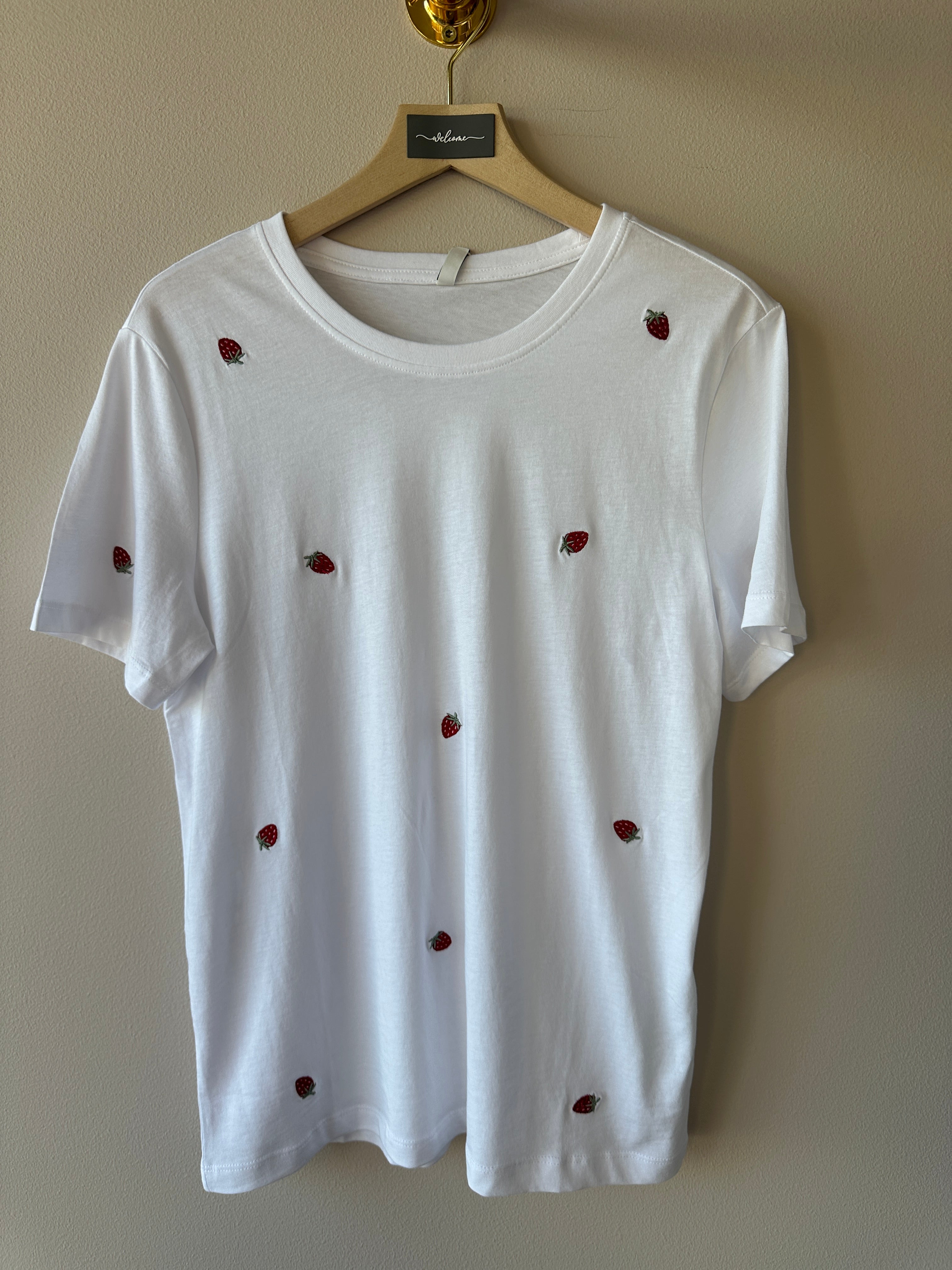 Vit oversized t-shirt med små broderade jordgubbar över hela tyget, rund hals och korta ärmar hängande på galge mot beige vägg.