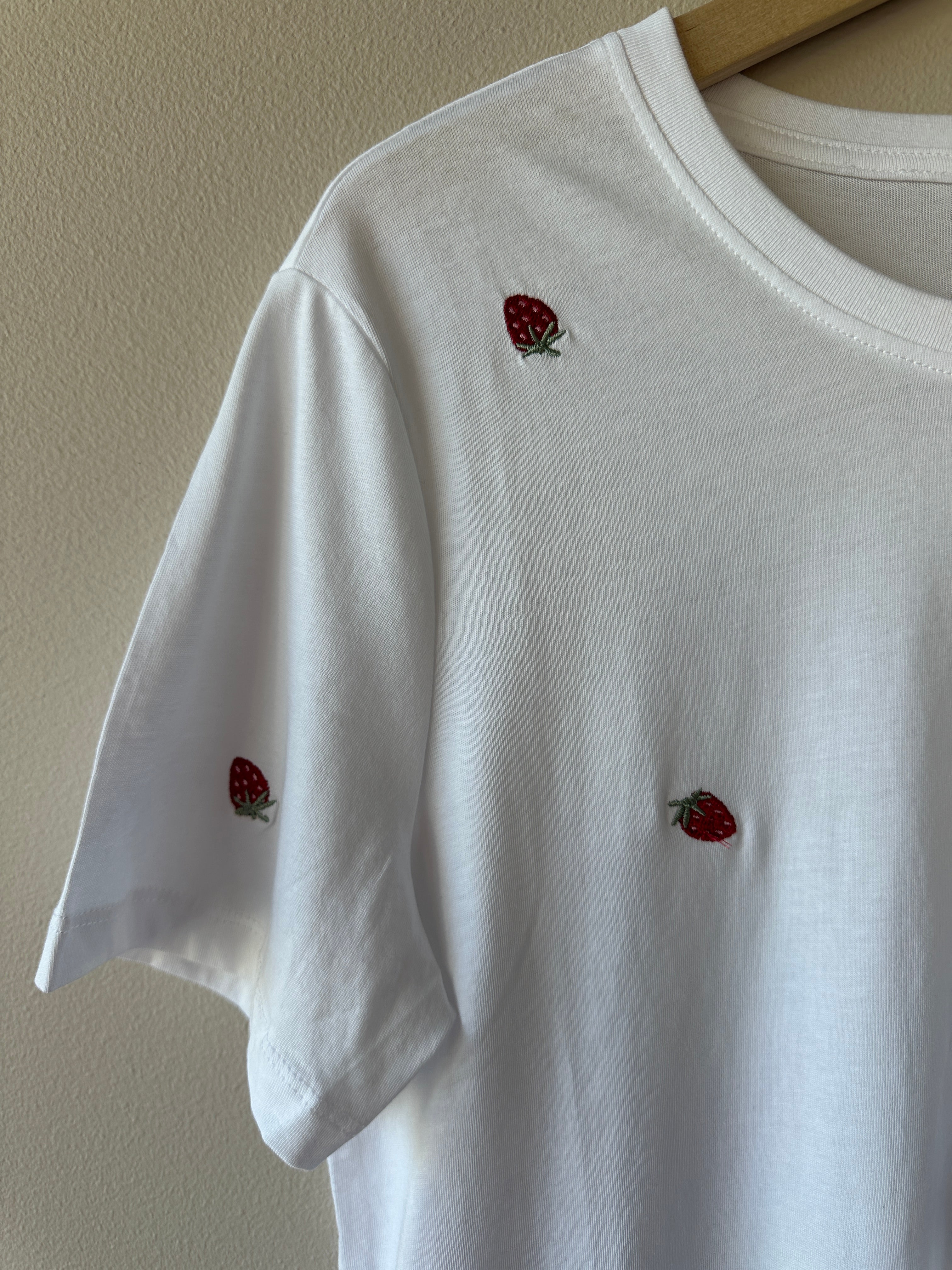 Närbild på vit t-shirt som visar broderade röda jordgubbar med gröna blad över tyget och ribbad rund hals, hängande på galge.
