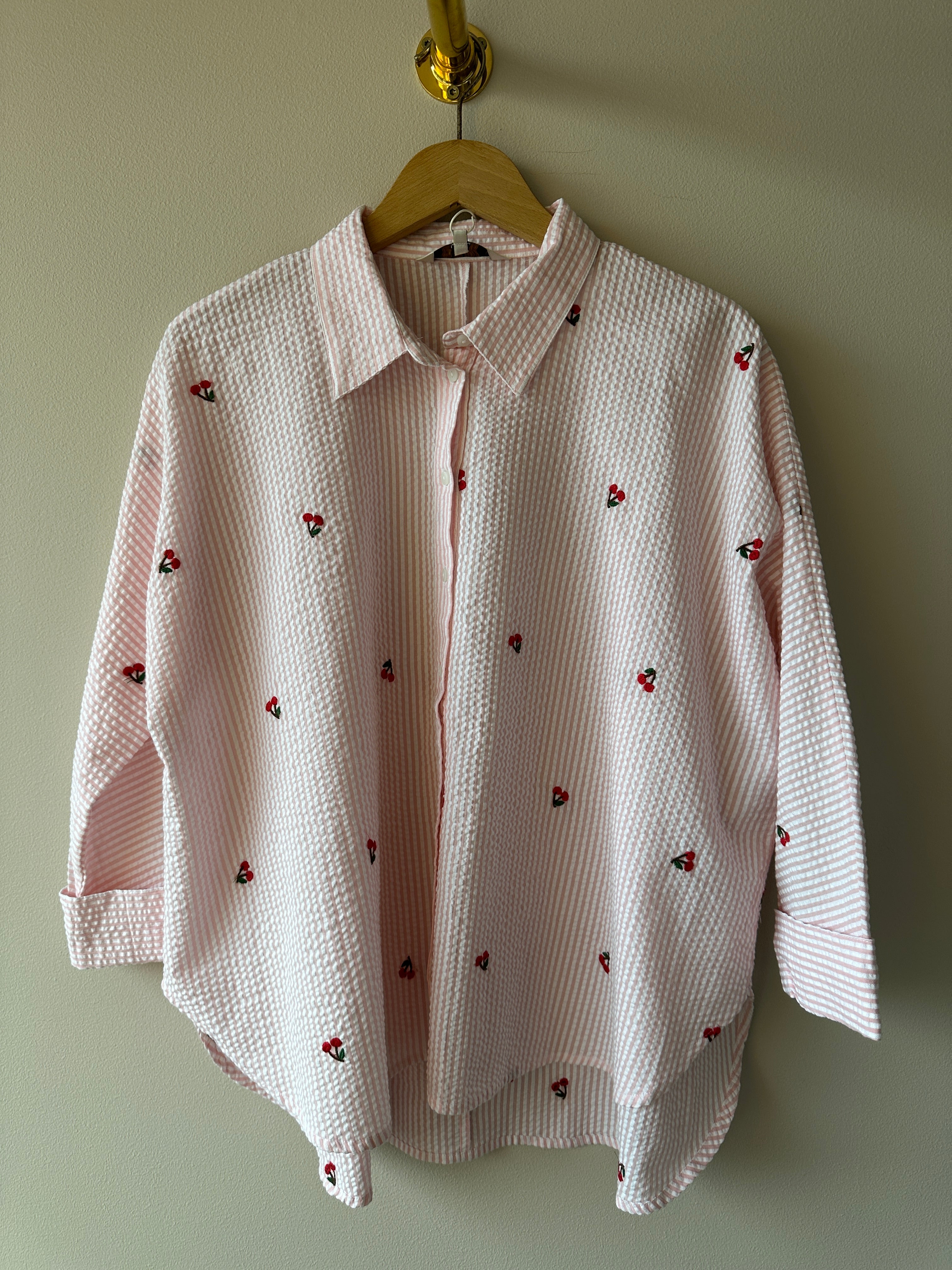 Vit seersucker-skjorta med rosa ränder och broderade röda körsbär, helbild framifrån hängd på galge mot gröngrå vägg.