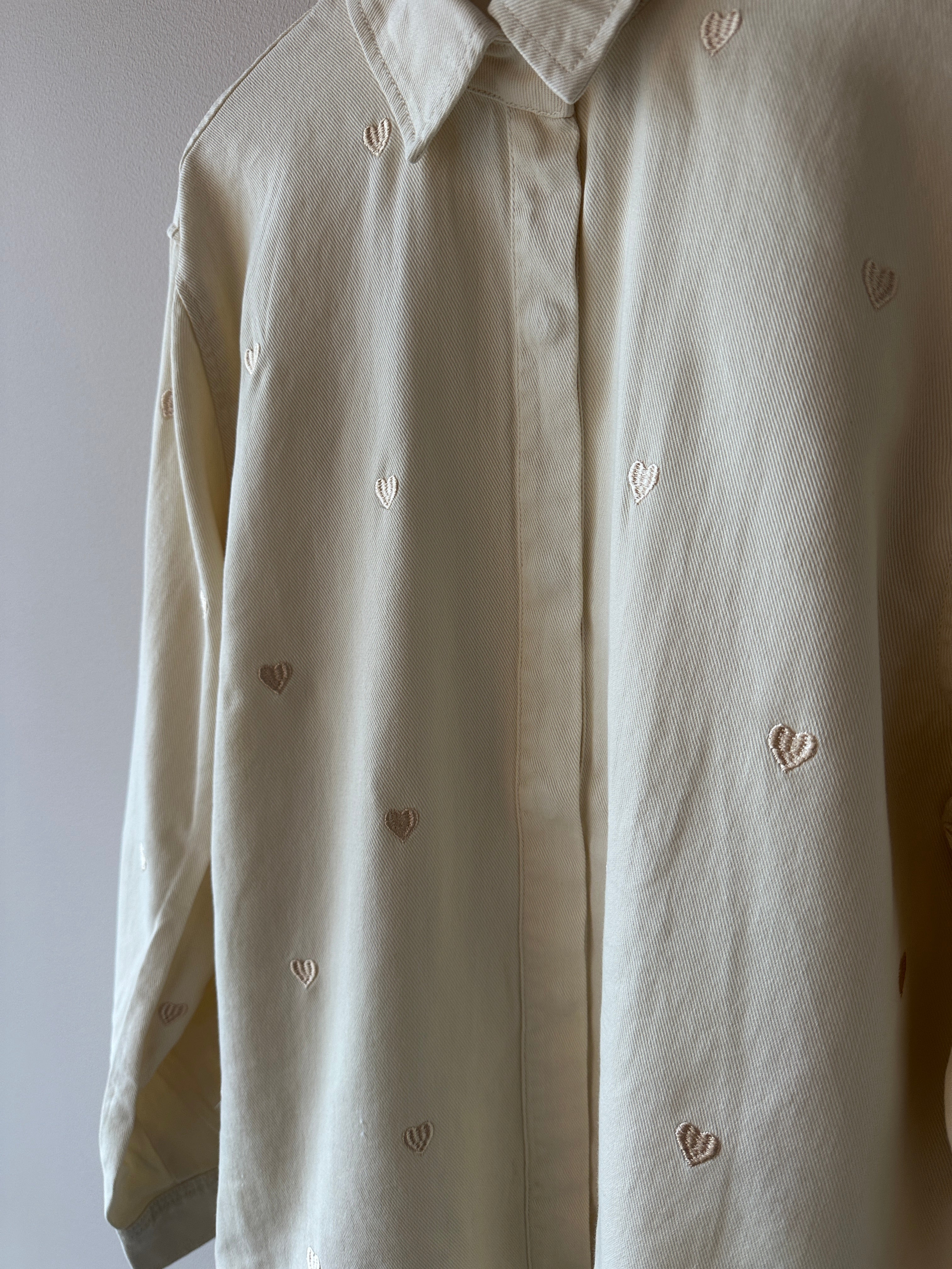 Närbild på crèmevit skjorta med broderade hjärtan över hela tyget och klassisk krage.