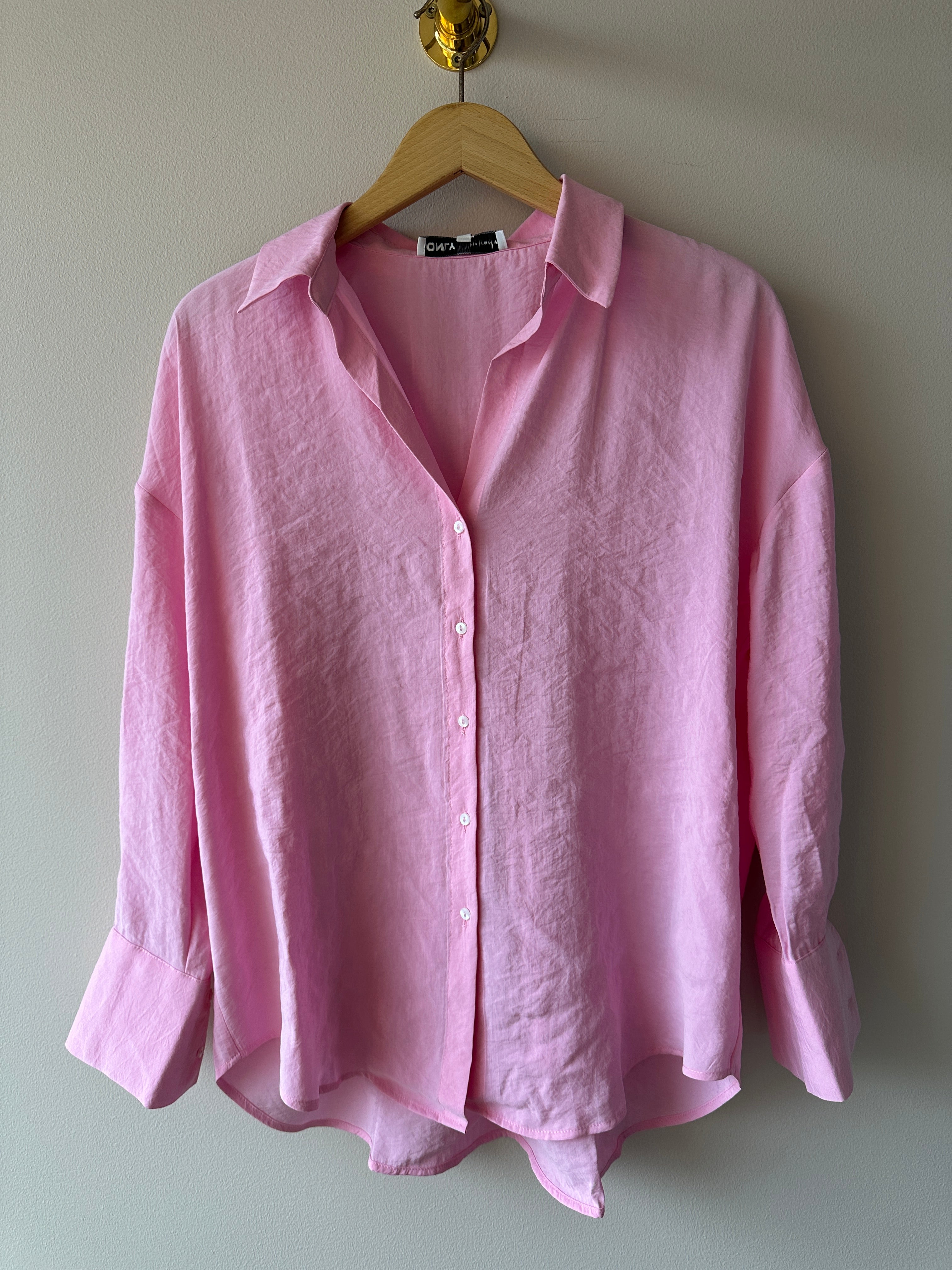 Rosa oversized linneskjorta med V-ringning, klassisk krage och vita knappar, hängd på galge mot ljusgrå vägg.