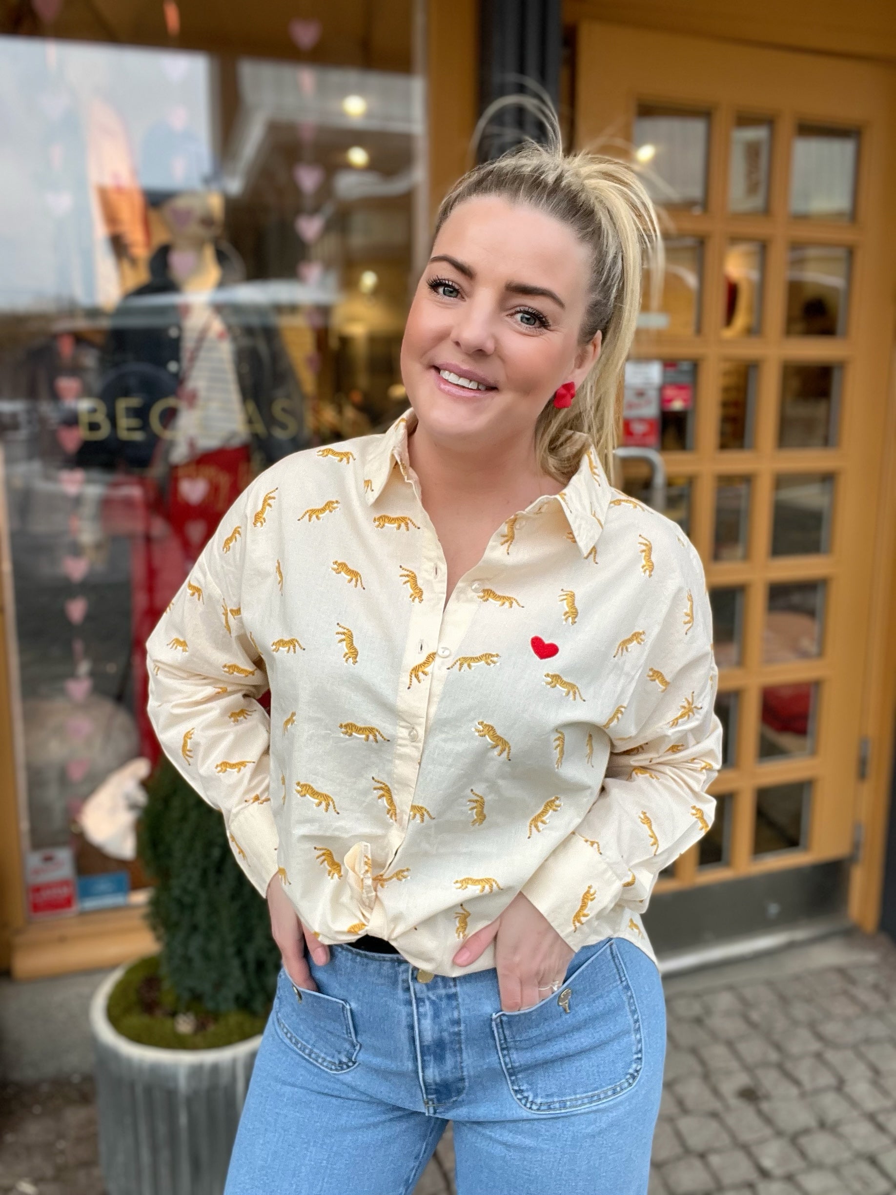 Leende kvinna framifrån med händerna i fickorna i cremevit oversized skjorta med guldigt tigertryck och litet rött hjärtbroderi på bröstet, stylade med ljusblå jeans