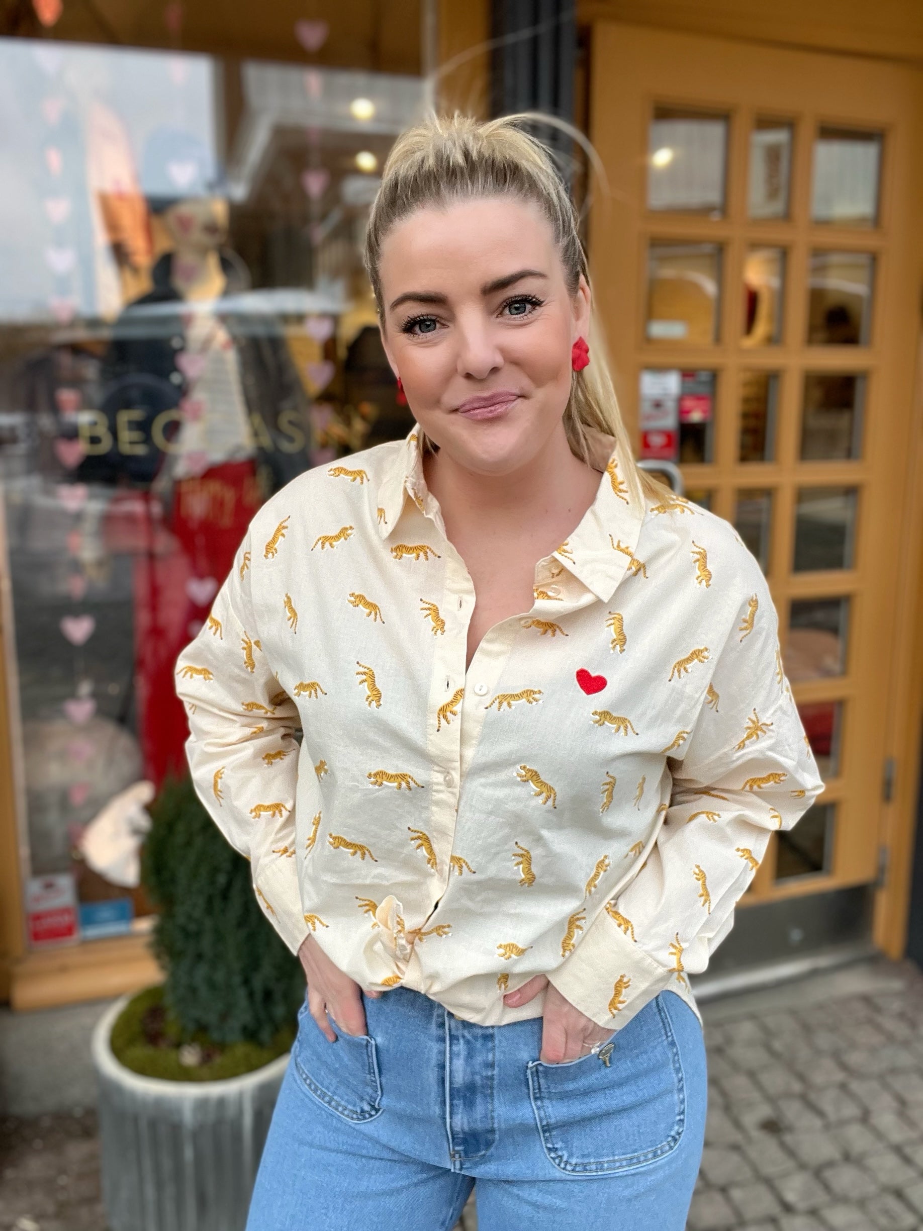 Leende kvinna med händerna i fickorna i cremevit oversized skjorta med tigertryck och rött hjärtbroderi på bröstet, stylade med ljusblå jeans och röda örhängen. 