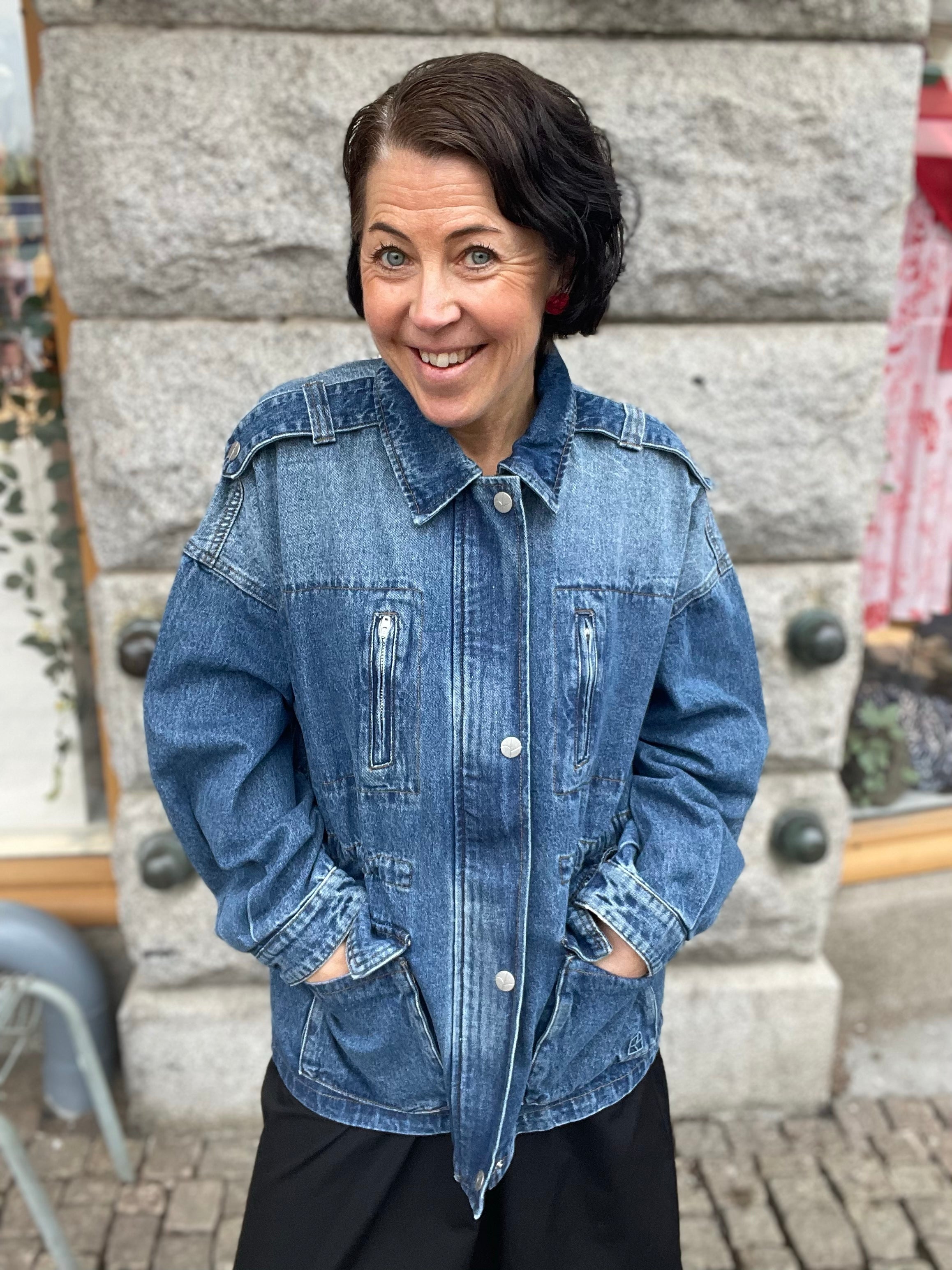 Leende kvinna framifrån i oversized mellanblå denimjacka med händerna i fickorna, fotograferad utomhus mot grå stenvägg.