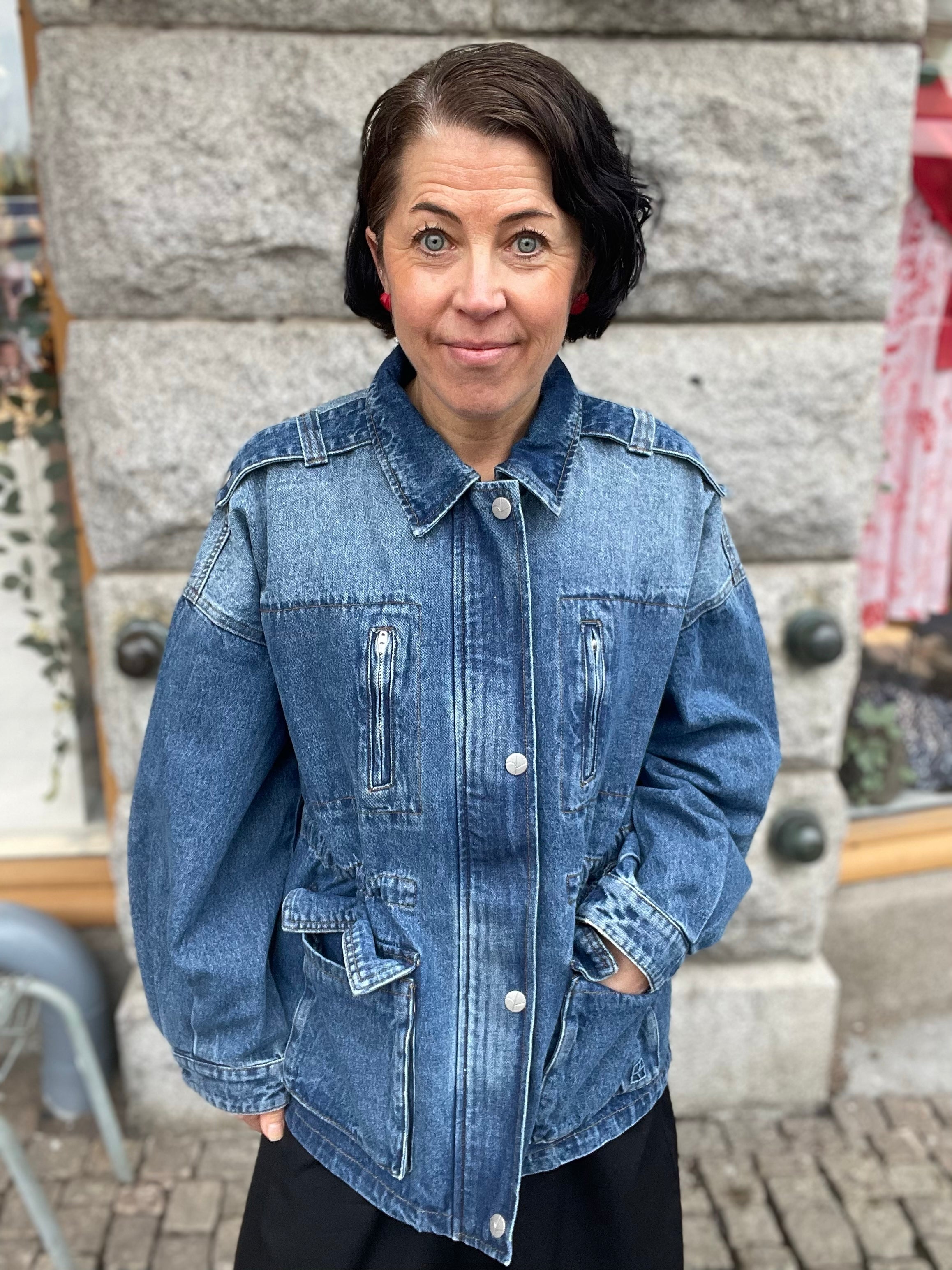Kvinna framifrån i oversized mellanblå denimjacka med klassisk krage, tryckknappar, dragkedjefickor på bröstet och stora patchfickor,  stylade med svarta byxor utomhus mot stenvägg.