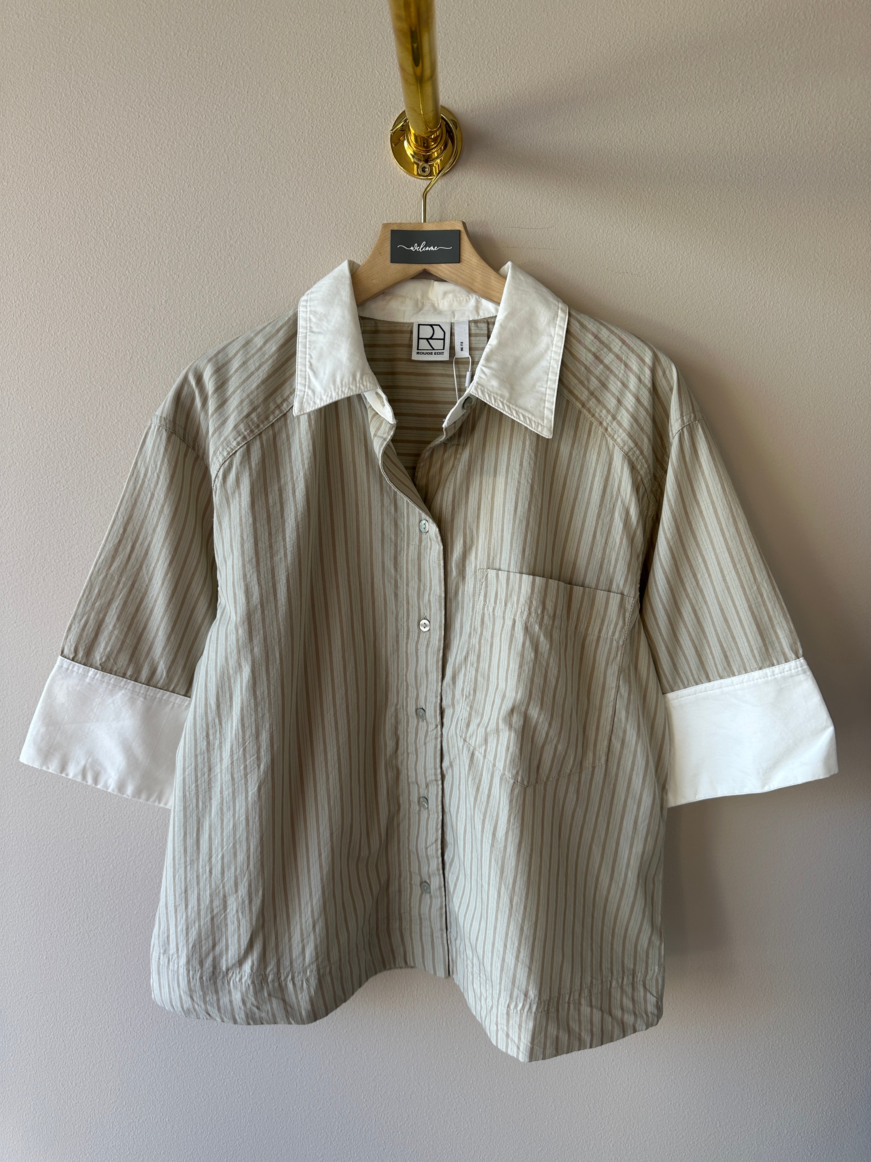 Beige och vit randig oversized skjorta med klassisk krage, korta ärmar med breda vita manschetter och brystficka hängande på galge mot beige vägg.