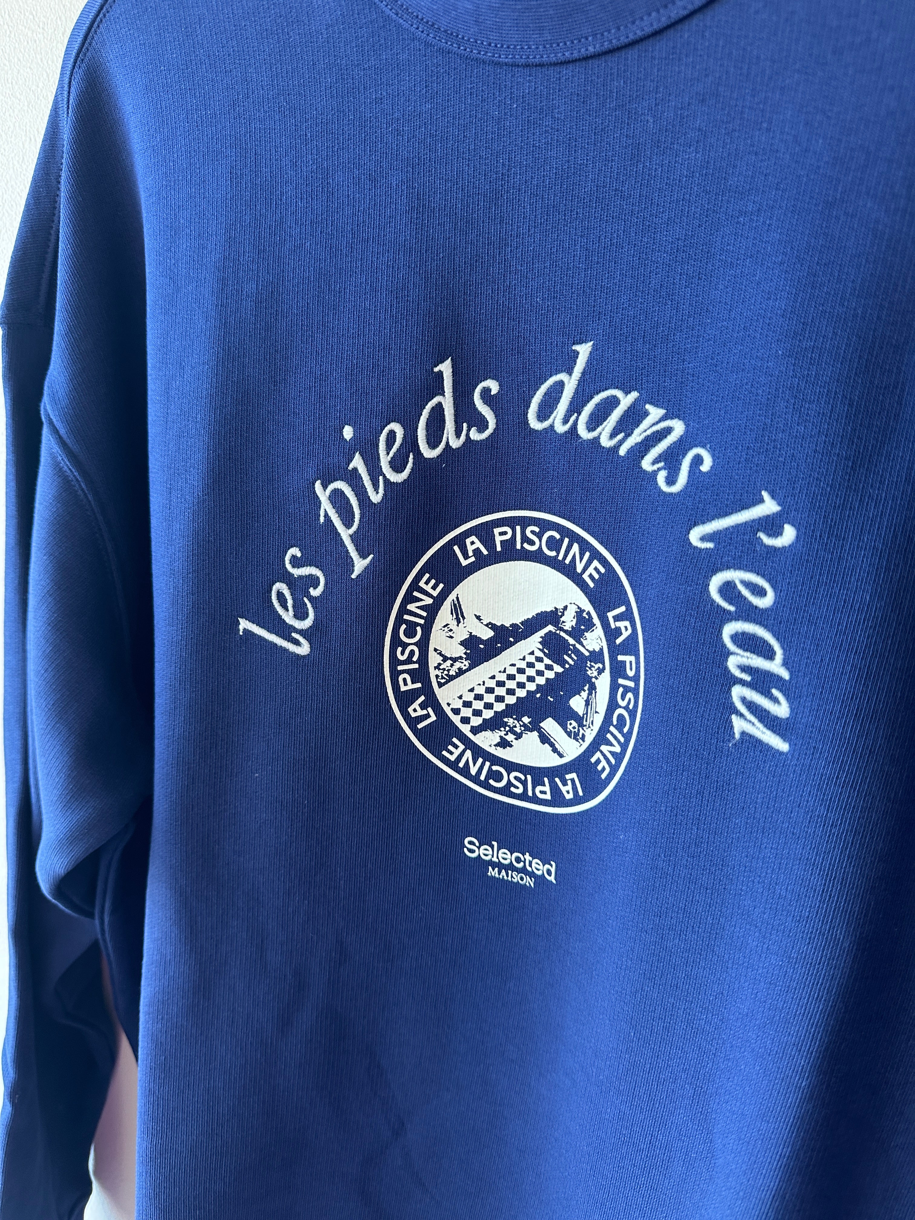Närbild på bröstet av marinblå sweatshirt som visar vitt kursivt tryck les pieds dans l'eau och runt La Piscine-emblem med poolmotiv samt Selected Maison-logotyp.