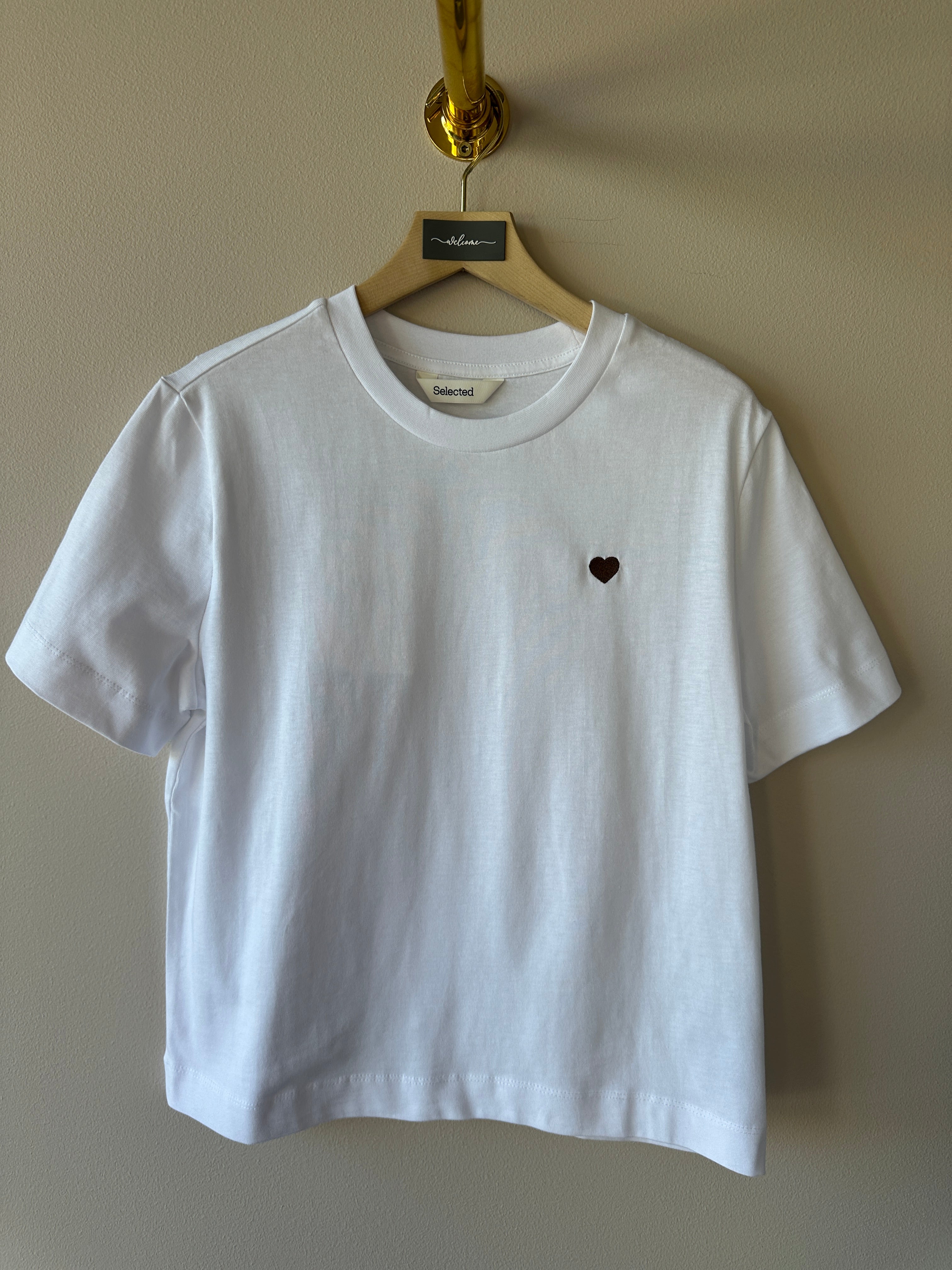 Vit oversized t-shirt med litet brunt hjärtbroderi på bröstet, rund hals och korta ärmar hängande på galge mot beige vägg, etikett med texten Selected synlig i halsen.