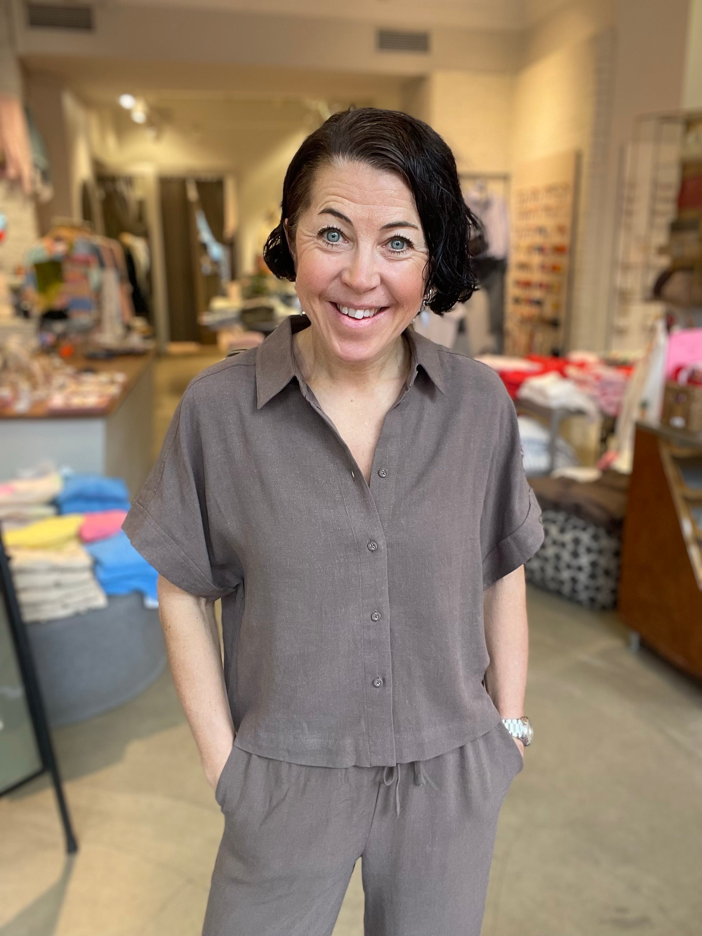 Leende kvinna framifrån i kortare gråbrun skjorta med klassisk krage och knappar, stylade med matchande vida byxor, fotograferad inomhus i butiksmiljö.