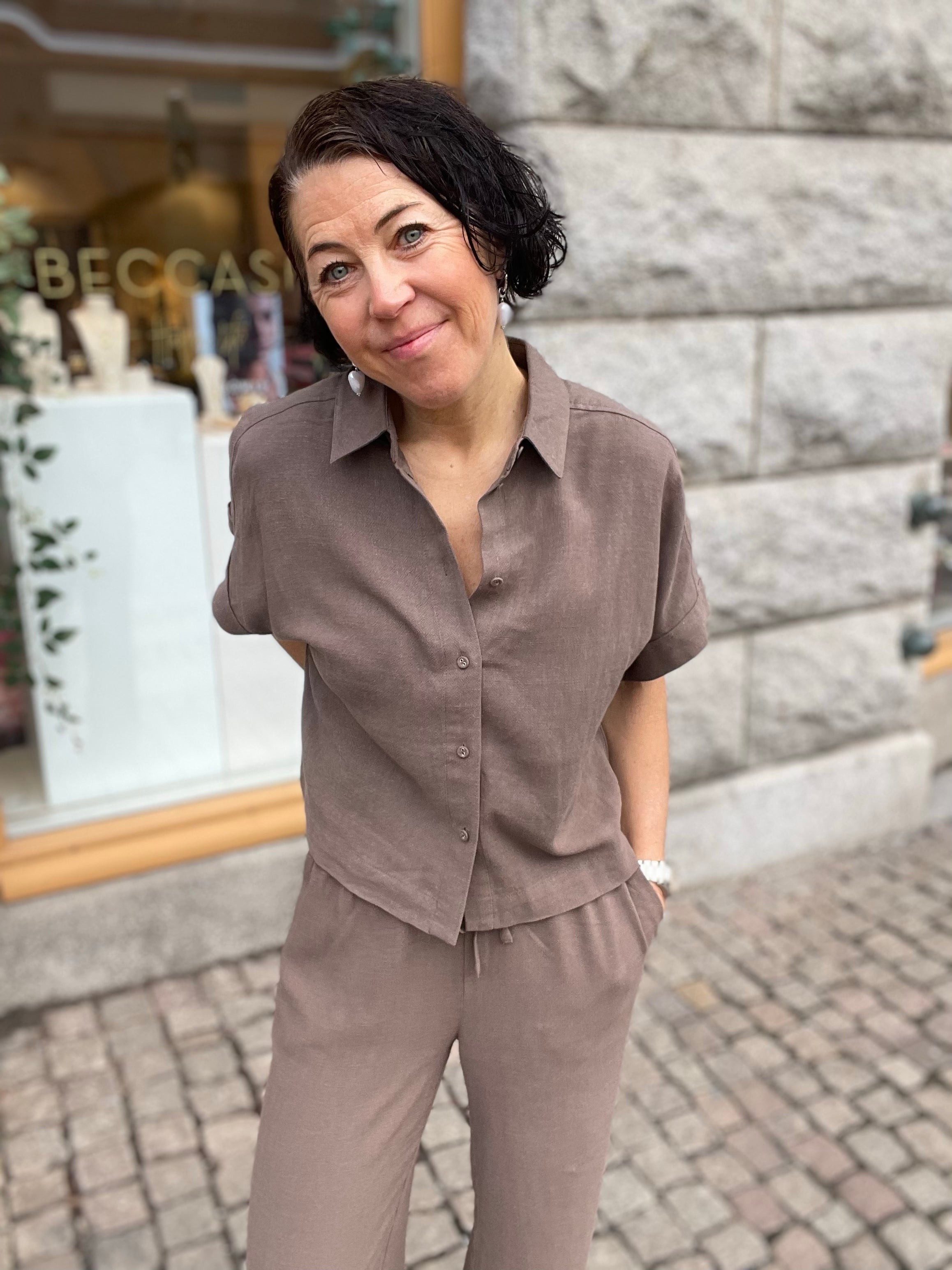 Kvinna i halvprofil i kortare gråbrun skjorta med klassisk krage och knappar, stylade med matchande vida byxor, leende utomhus mot grå stenvägg med butiksskylt i bakgrunden.