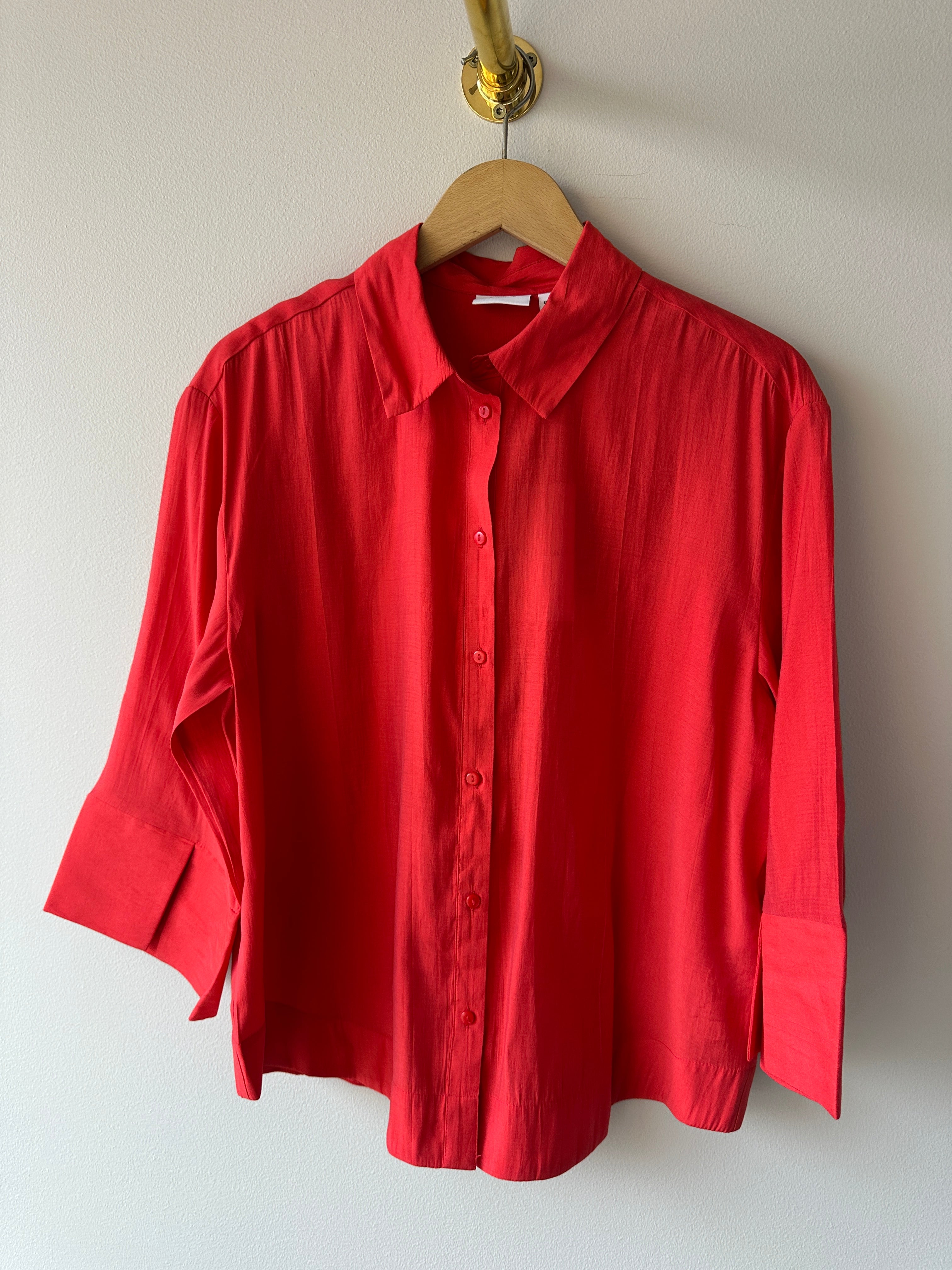 Klarröd oversized skjorta med klassisk krage, matchande knappar och 3/4-ärmar i lätt flytande tyg, hängd på galge mot vit vägg.