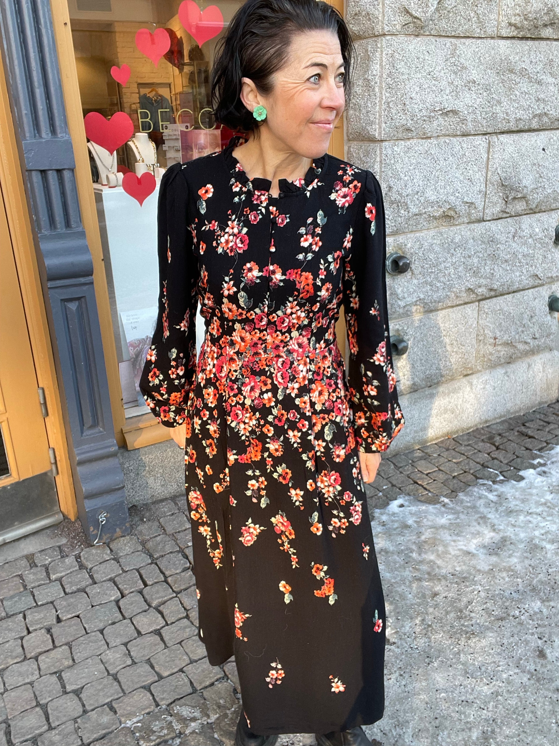 Kvinna i halvprofil i lång svart klänning med rött och orange blomtryck, volangkrage vid halsen och långa puffärmar. Fotograferad utomhus framför butik med hjärtdekorationer i fönstret.