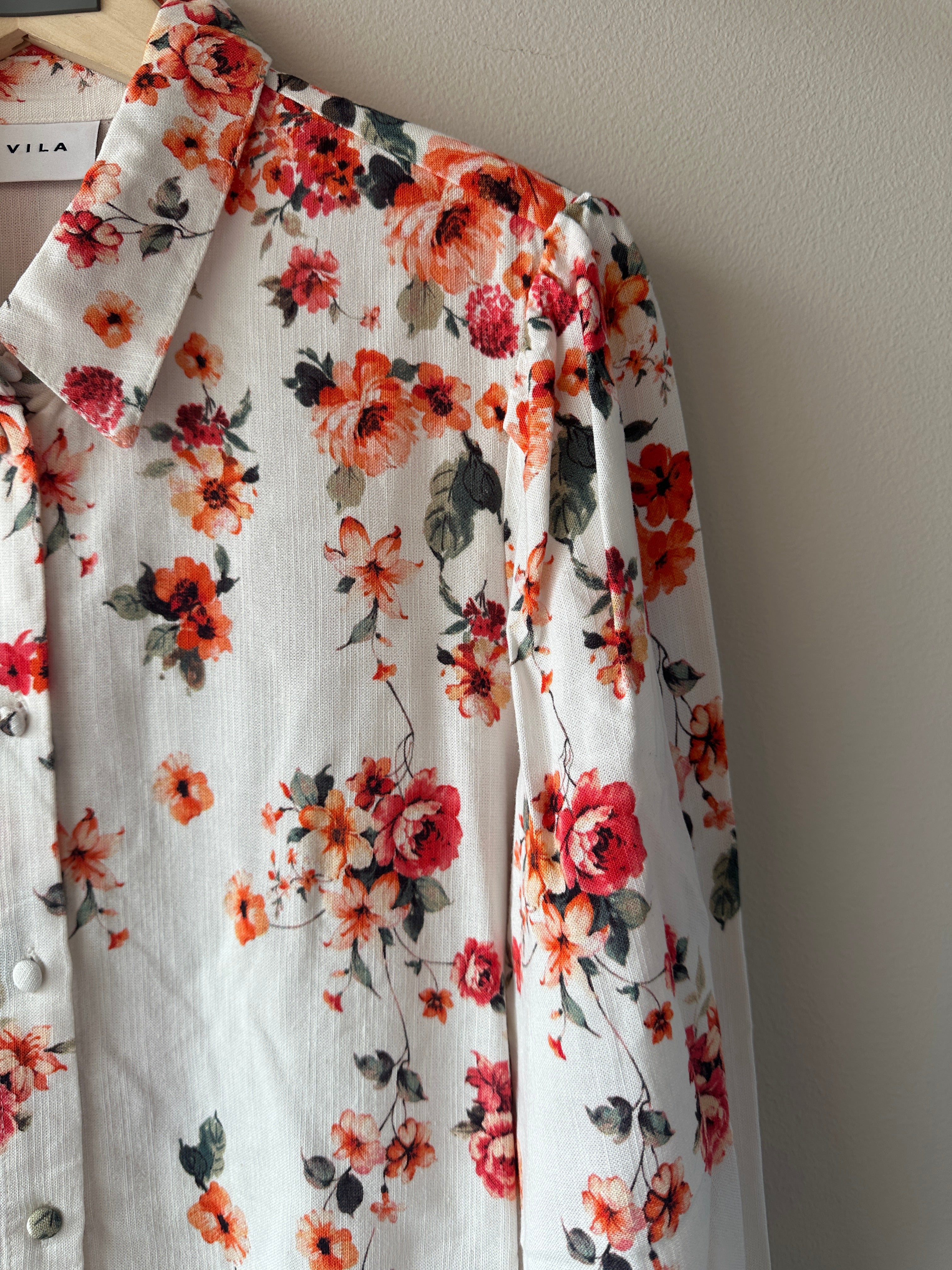 Vit skjorta med rött och orange blommigt tryck, klassisk krage, knappar fram och långa puffiga ärmar med smockade manschetter, hängande på galge mot beige vägg, etikett Vila synlig i halsen.