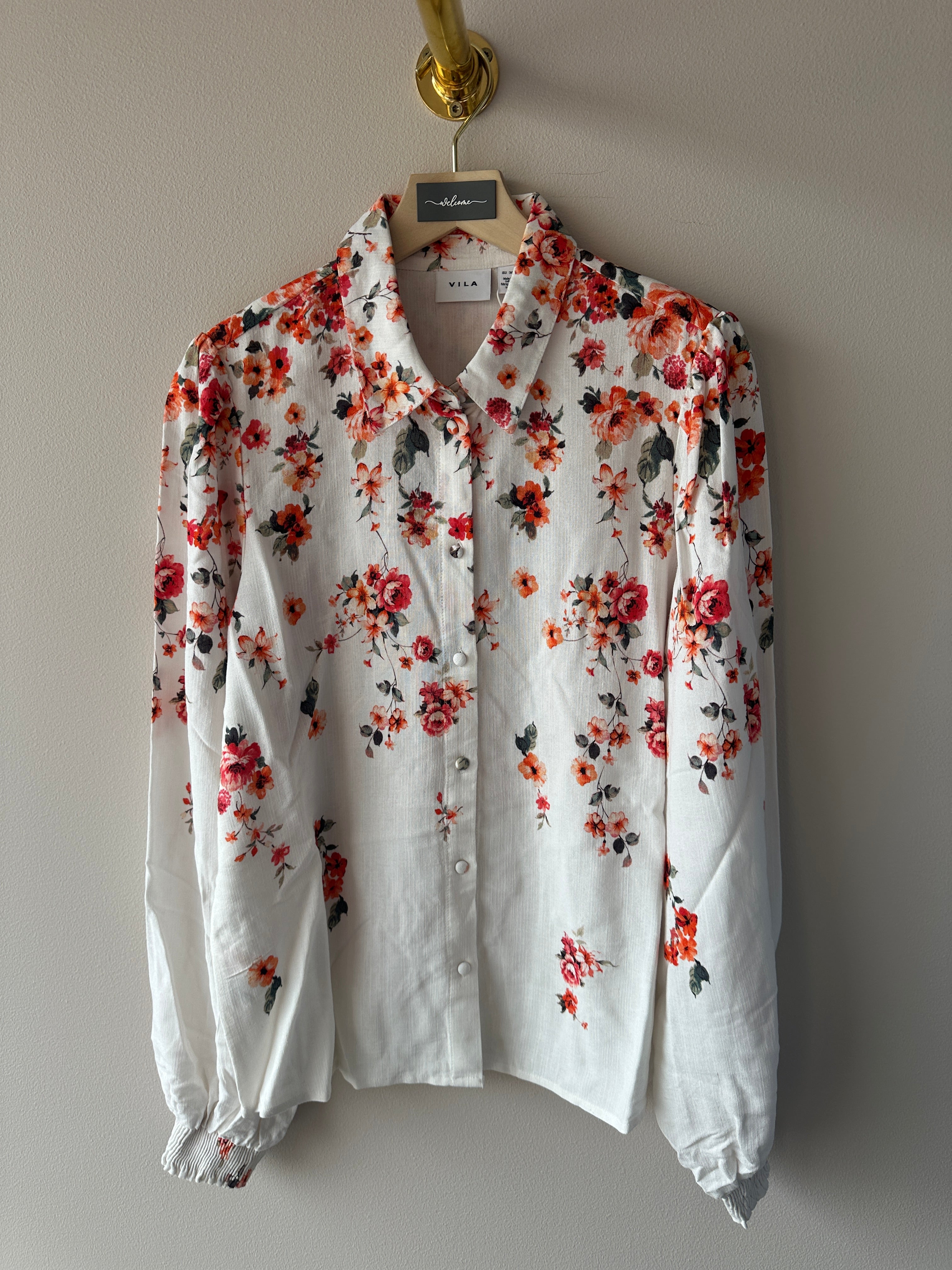 Närbild på övre del av vit skjorta med rött och orange blommigt tryck i varierande storlekar, klassisk krage och knappar fram, hängande på galge, etikett Vila synlig.